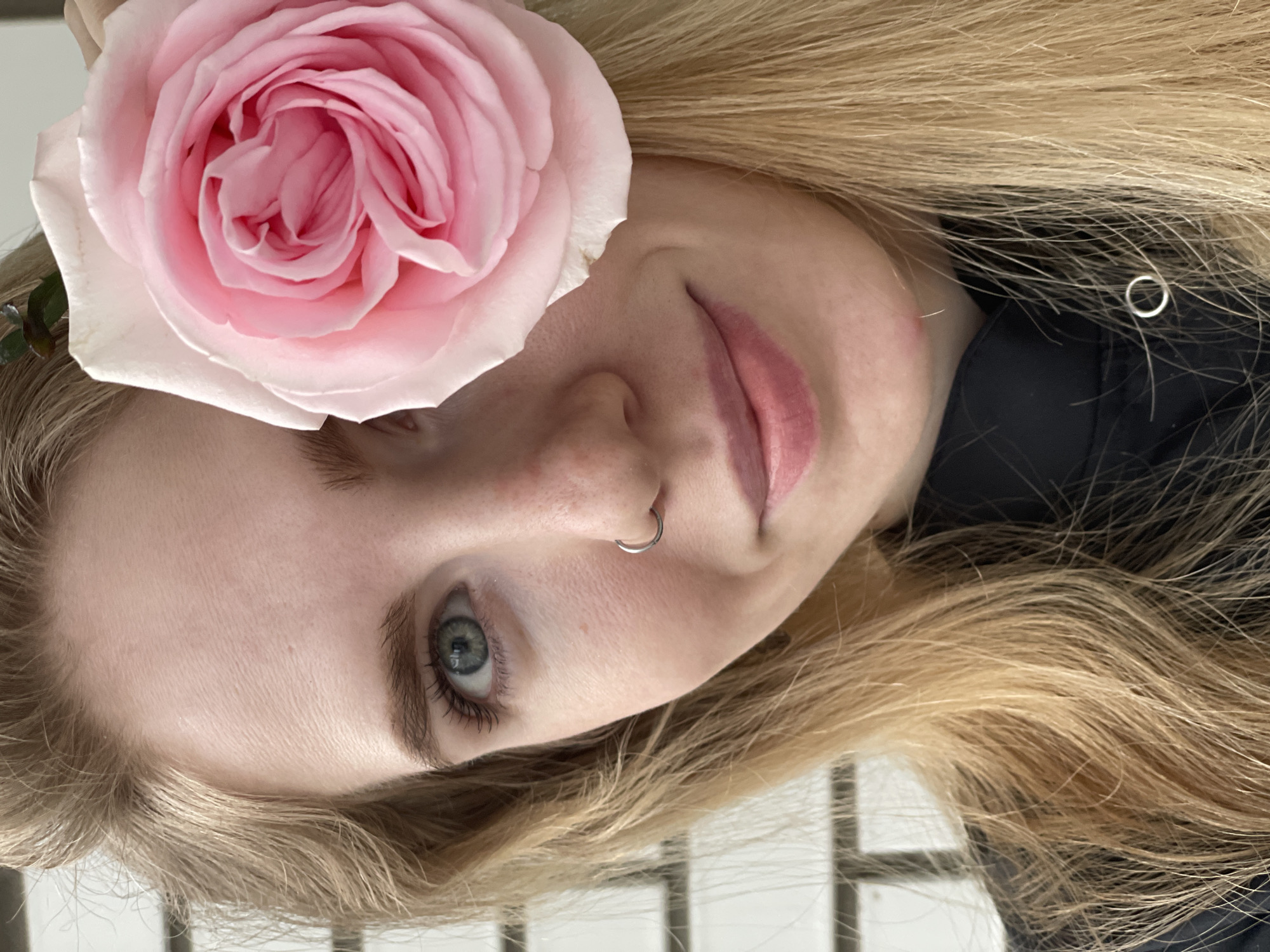 A close-up image features a woman's face, turned slightly to the side. Her hair is light blonde and wavy. A large pink rose adorns her head, positioned above her forehead. She has fair skin, blue eyes, a nose ring, and pink lips. A dark-colored garment is visible around her neck. The background includes what appears to be a blurred window or a tiled surface. The scene does not contain any indicators of Izhevsk, Russia, and cannot be identified to be in that location.