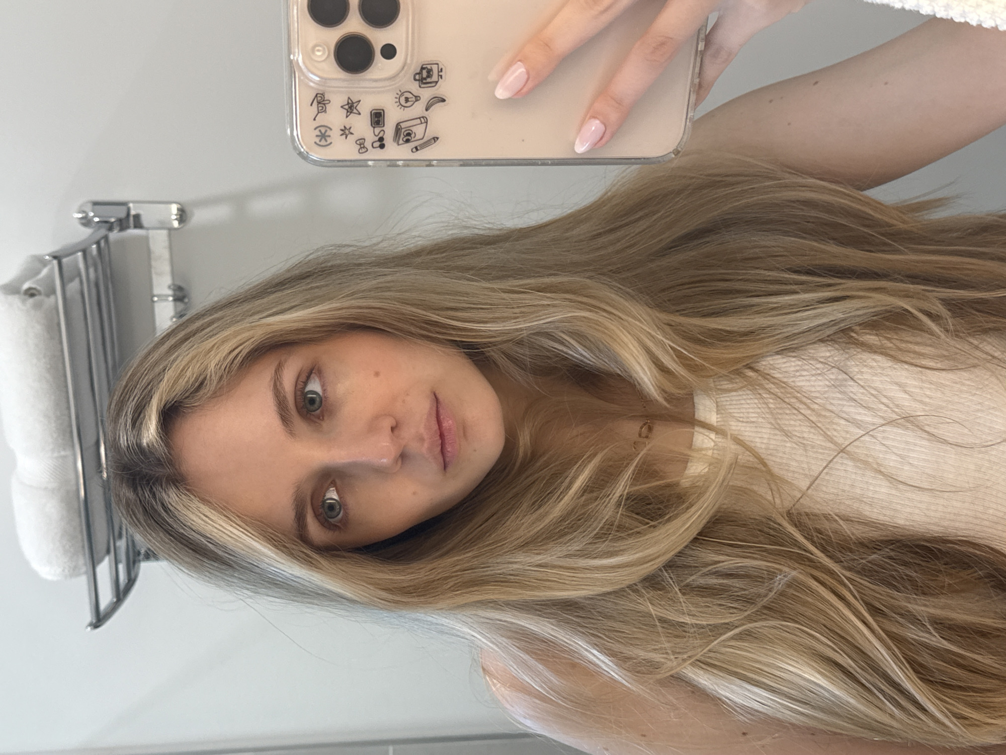 In Miami, Florida, a young woman with long blonde hair takes a mirror selfie in a bathroom. She is wearing a white tank top and has a delicate necklace. Her fingernails are painted a soft pink. The background shows a bathroom shelf with folded white towels and a chrome rack. The woman is holding a clear phone case with various black doodle stickers, including a book, a lightbulb, and musical notes.