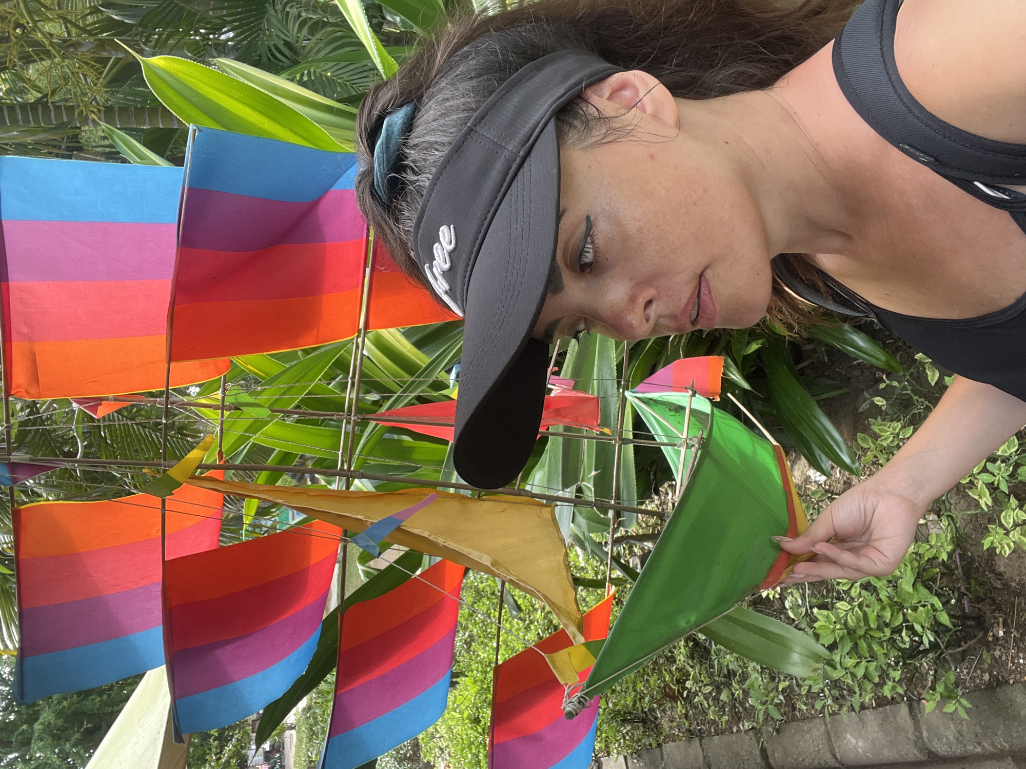 A woman wearing a black visor with the word "FREE" embroidered in white is leaning over a colorful display of kites. The kites are made of fabric and have horizontal stripes in various vibrant colors, including orange, red, purple, and blue. Some kites are shaped like sails, while others are rectangular. The woman is holding a green kite in her right hand, appearing to adjust it. Her face is partially obscured by the visor, but she has dark hair pulled back and is looking down at the kites with a focused expression. The background consists of lush green foliage, suggesting an outdoor setting. The lighting indicates it is daytime, with sunlight filtering through the leaves. The scene is likely taking place in Kuta Selatan, Indonesia.