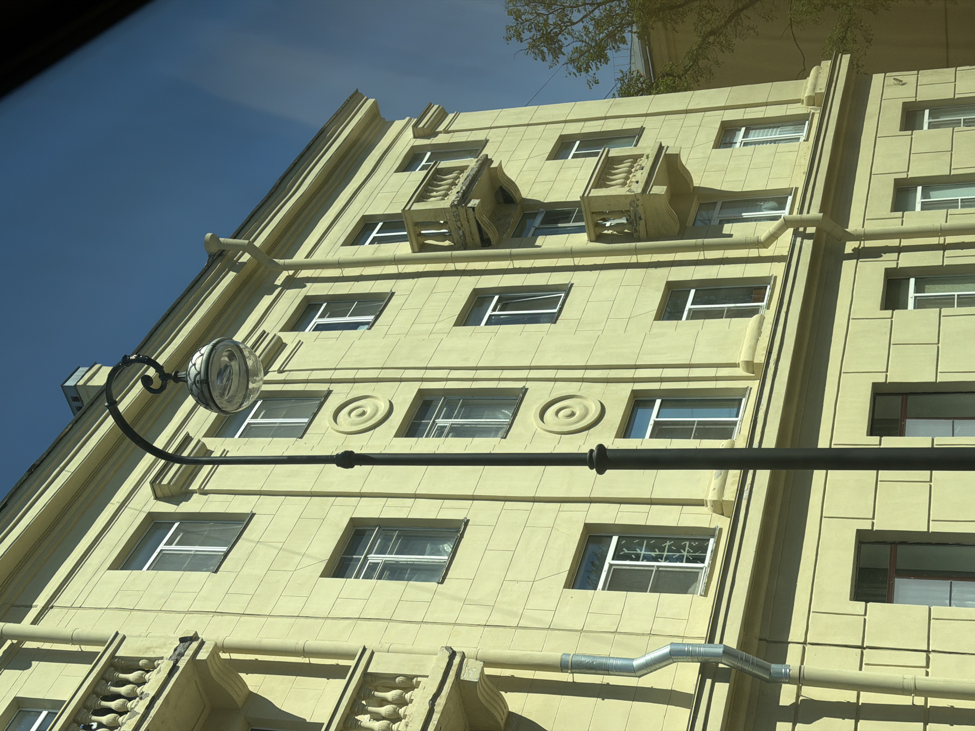 The image shows the facade of a yellow multi-story building in Kronstadt, Russia. The building has evenly spaced windows on each floor. Decorative elements such as circular designs and ornate balconies embellish the facade. A black street lamp and a metallic pipe run horizontally across the building's front. The sky above is blue, with tree branches visible in the upper part of the image.