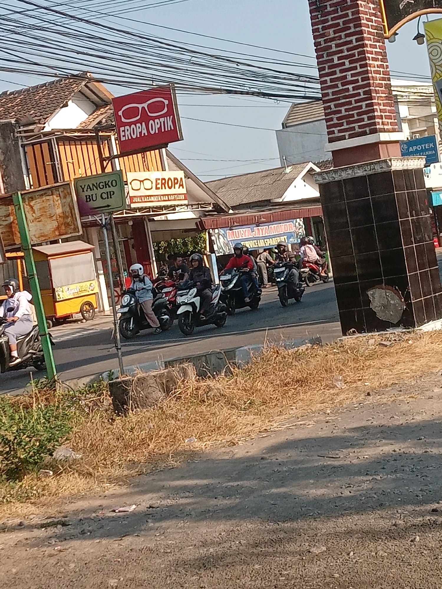 This is a street-level photograph taken in Banguntapan, Indonesia, on a sunny day with a clear blue sky. The scene captures a busy intersection with several motorcycles on the road and pedestrians on the sidewalk.
On the left side of the image, a red sign with white glasses prominently displays "EROPA OPTIK," indicating an optical shop. Below it, a green sign reads "YANGKO BU CIP," and another sign for "EROPA" with glasses is visible, stating "JUAL KACAMATA DAN SOFTLENS." Further to the right, a brick pillar marks the entrance to a business.
Several people are riding motorcycles, some wearing helmets. One person on a white motorcycle is on the far left, followed by others on dark-colored motorcycles. In the background, buildings line the street, including what appears to be "MULYA JAYA MOTOR" with a banner that reads "SERVICE DAN SPAREPART."
The road is paved, and the sides are lined with dry grass and some greenery. The overall impression is of a typical commercial street scene in a populated area.