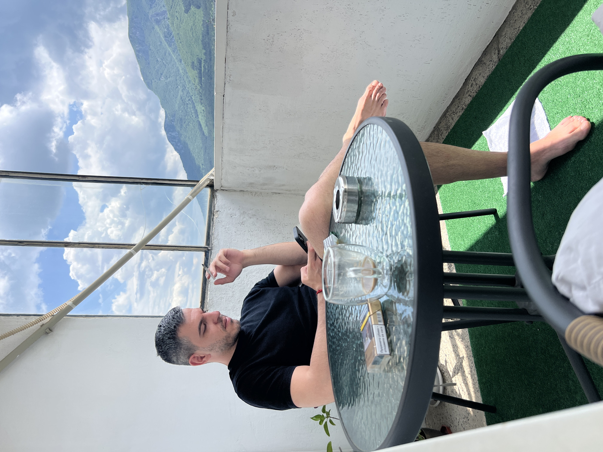 A man, appearing to be in his late twenties to early thirties with short, dark hair showing some grey, is seated on a balcony in Vratsa, Bulgaria, during daytime. He wears a black short-sleeved t-shirt and has bare feet; one foot rests on a small white towel placed on a green artificial turf floor. He holds a lit cigarette in his right hand and a smartphone in his left, which he is looking down at. A red string bracelet is visible on his left wrist.
He sits at a round glass-top table with a dark metal frame and legs. On the table, a gold-colored cigarette pack displaying the Bulgarian health warning "ПУШЕНЕТО УБИВА – ОТКАЖЕТЕ СЕ СЕГА" (SMOKING KILLS – QUIT NOW) is present, along with a clear, double-walled glass mug containing a small amount of light brown liquid, and a metallic, circular ashtray.
The balcony features white, textured walls. A large window with dark frames and a horizontal support bar is visible to the left and behind the man. To the right, beyond a low balcony wall, a panoramic landscape of lush green mountains stretches under a bright blue sky dotted with scattered white clouds, consistent with the geography surrounding Vratsa. A small potted plant with green leaves is visible on the far left of the balcony. The scene is illuminated by natural sunlight, casting shadows.
