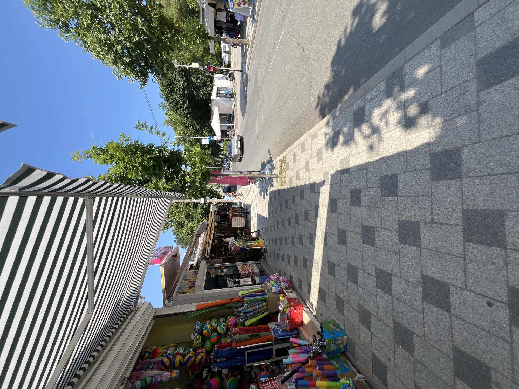 The image depicts a street scene in Obzor, Bulgaria, on a sunny day. On the left side of the frame, a storefront is visible, adorned with a black and white striped awning. The store displays various items, including colorful balls, umbrellas, and other beach-related merchandise, arranged on the sidewalk and within the shop. The sidewalk is paved with grey tiles, some of which are darker, creating a patterned effect. Shadows of trees are cast across the sidewalk.
The street itself is paved with asphalt and appears to be moderately busy. A silver car is driving on the road, and a white tent structure with tables and chairs is set up on the opposite side of the street. Further down the street, more shops with awnings are visible, and several people are walking. The environment is characterized by trees lining the street and a clear blue sky.
