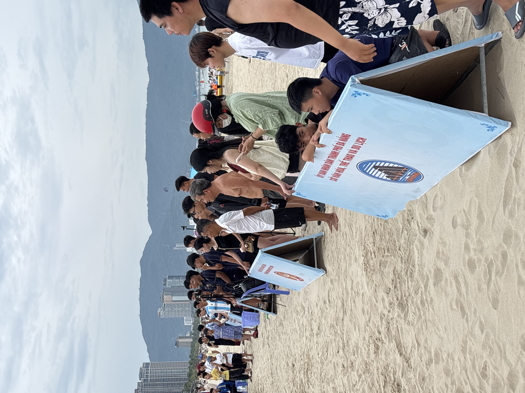A group of people, some wearing sportswear, stand on a sandy beach facing left, where a body of water is likely located. In the foreground, two large, light blue triangular signs with red and blue graphics and Vietnamese text are propped up on the sand. The text on one sign reads "ỦY BAN NHÂN DÂN THÀNH PHỐ ĐÀ NẴNG SỞ VĂN HÓA, THỂ THAO VÀ DU LỊCH," which translates to "PEOPLE'S COMMITTEE OF DA NANG CITY DEPARTMENT OF CULTURE, SPORTS AND TOURISM." In the background, buildings and mountains are visible under a cloudy sky. Several individuals are wearing athletic attire, and some appear to be participants or spectators at an event. One person wears a red helmet and a face mask. The overall scene suggests an outdoor event or competition taking place on a beach in Da Nang, Vietnam.