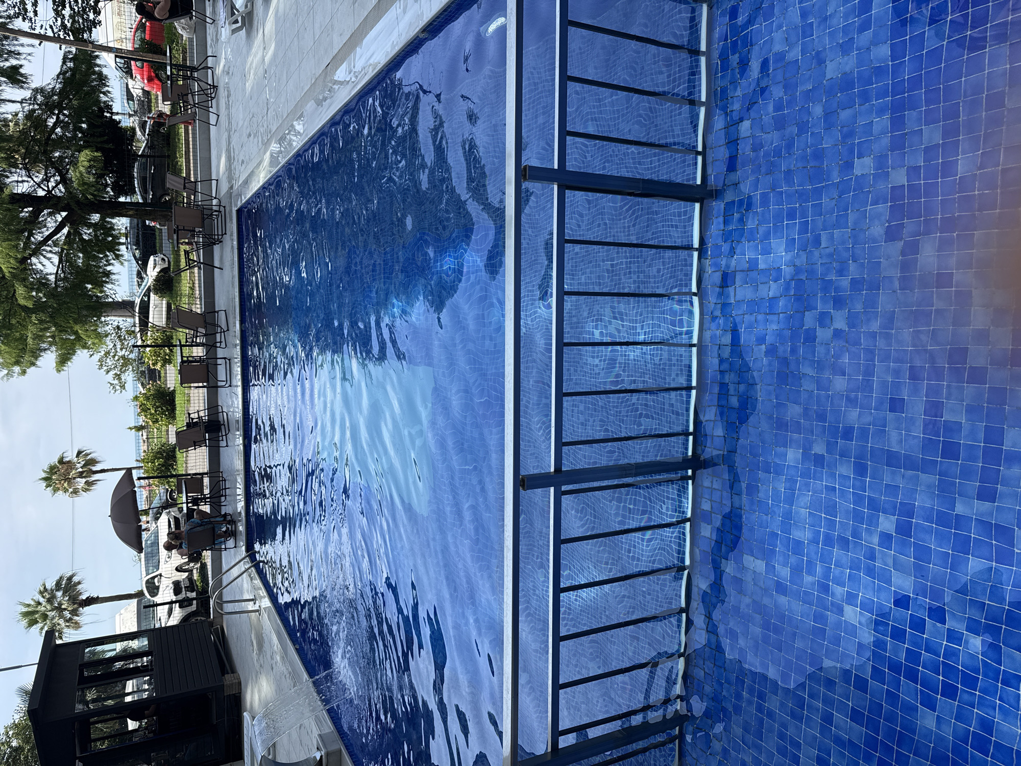 The image depicts an outdoor swimming pool at a facility near the sea in Batumi, Georgia. The pool is filled with blue, tiled water and has a metal railing in the foreground. In the background, several cars are parked, and a few palm trees and other trees are visible. There are also tables and chairs set up outside, suggesting an area for dining or relaxation. A dark-colored, modern-looking structure with windows, possibly a bar or reception area, is located to the left of the frame. The sky is overcast, indicating a cloudy day. The overall scene suggests a resort or hotel setting.