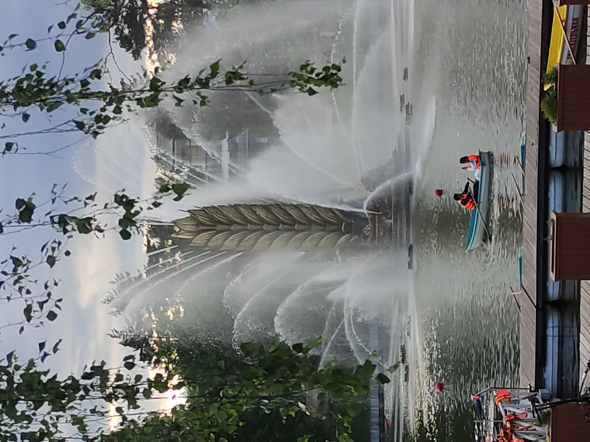 Here's a detailed description of the image:
**Scene Overview:** The image captures a vibrant urban scene featuring a large, ornate fountain in the middle of a body of water, likely a pond or small lake. Two people in a rowboat are positioned near the fountain. Lush greenery frames the scene, creating a park-like atmosphere.
**Subjects & Objects:**
* **Fountain:** The fountain is the central element, characterized by a decorative structure that resembles a giant leaf or a plume of feathers. Water shoots out in radial patterns, adding to the dynamism.
* **Rowboat:** A small rowboat with two occupants wearing life vests is visible near the fountain.
* **People:** Two people in the boat, appear to be wearing orange life vests.
* **Vegetation:** Trees and bushes surround the water, with some branches partially obscuring the view.
* **Dock/Boardwalk:** A dock or boardwalk runs along the edge of the water body, with some infrastructure elements visible.
**Actions & Interactions:**
* The fountain is actively shooting water in various directions.
* The two individuals in the rowboat are seemingly navigating near the fountain.
**Environment:**
* The scene is set in an urban park.
* The weather appears to be clear, with sunlight filtering through the trees.
* The water in the pond is reflective.
**Location:** The location in the city of Unknown, Unknown.