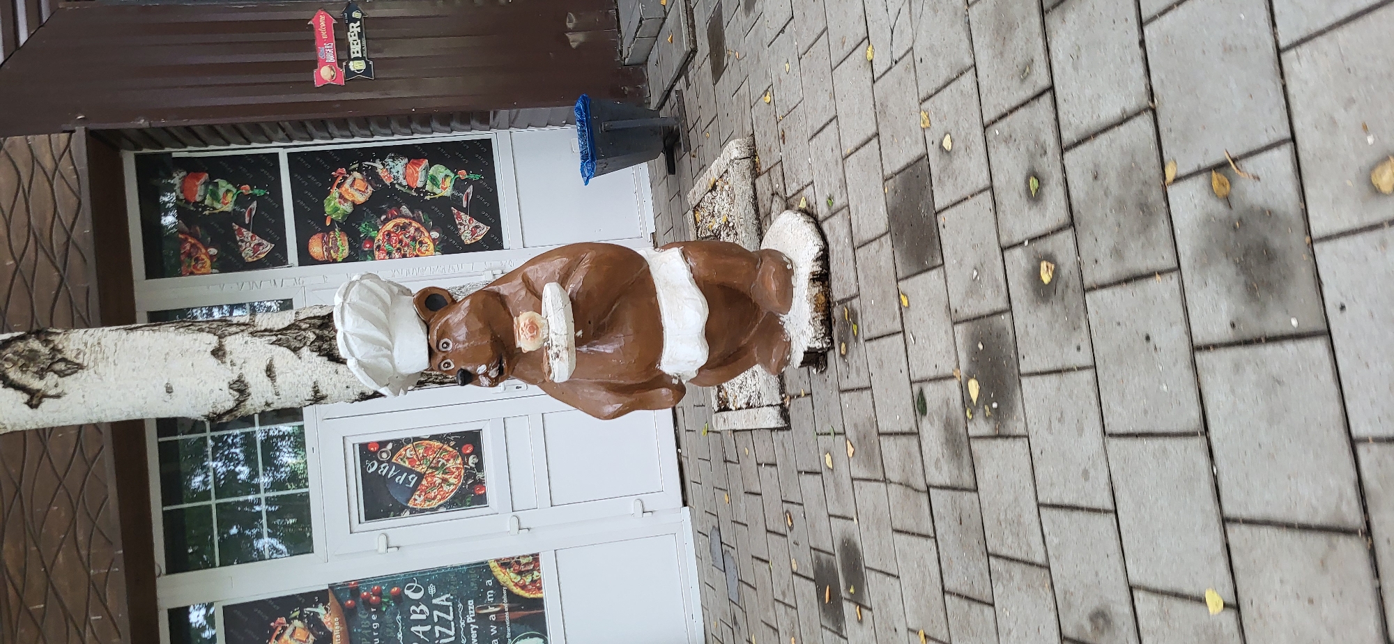The image shows a statue of a brown bear wearing a chef's hat and apron, holding a plate with a small pastry on it. The statue is placed on a sidewalk made of gray rectangular pavers, with scattered yellow leaves on the ground. In the background, there is a building with white doors and windows decorated with posters of pizza, burgers, and sushi. There is also a brown wooden wall on the right side of the image with two directional signs, one pointing left with the word "BURGERS" and "welcome", and another pointing left with the word "BEER". A blue trash can is visible near the brown wall. The overall scene appears to be in front of a cafe or restaurant. The location is in the city of Oskemen, Kazakhstan.