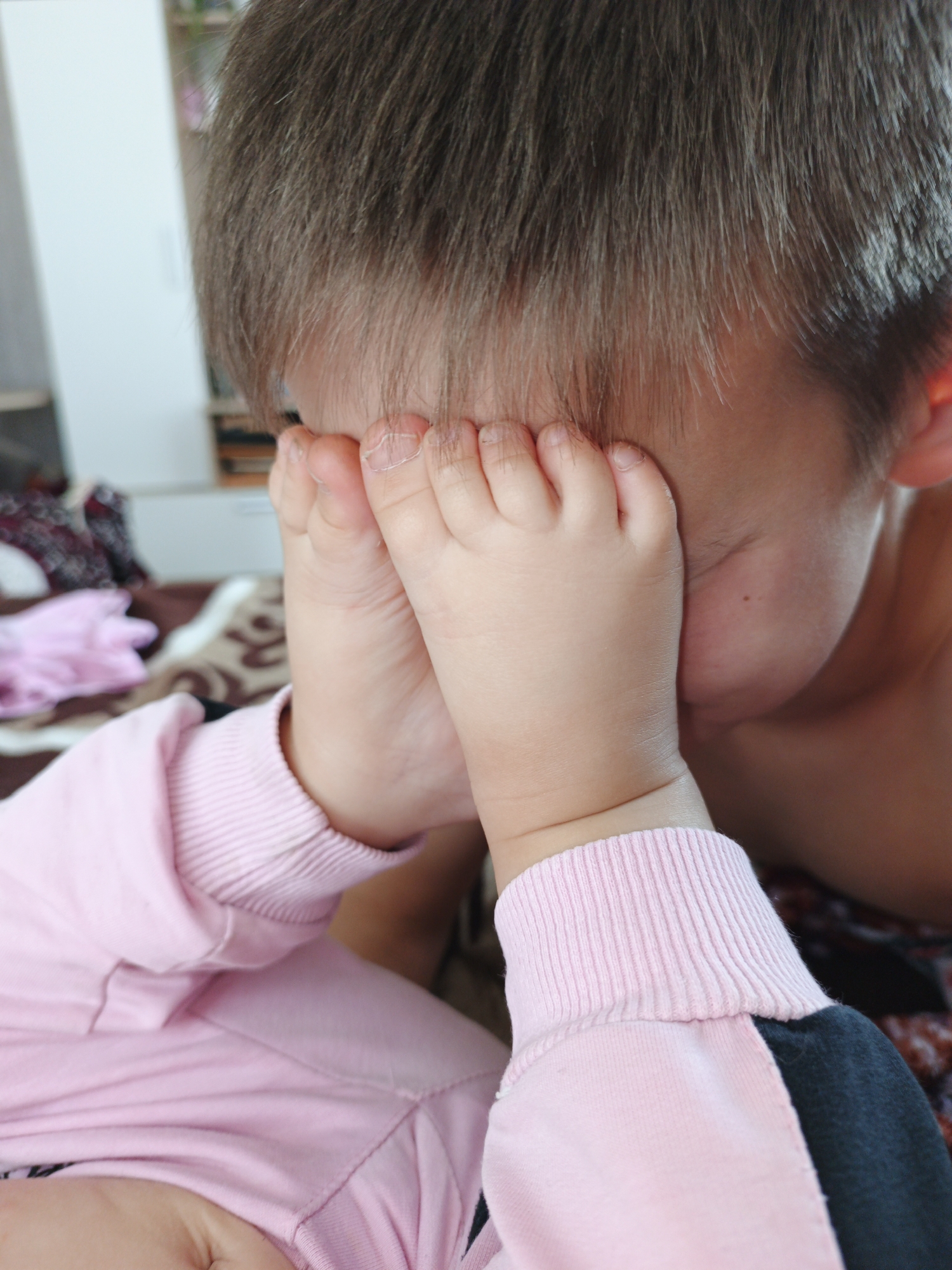 The image is a close-up, eye-level shot of a young child covering their face with their feet. The child has brown hair and is wearing a pink sweatshirt with a dark sleeve. The background is blurred but appears to be an indoor setting with furniture and bedding. There is no visible indication of the location being Medvedyovka, Russia.