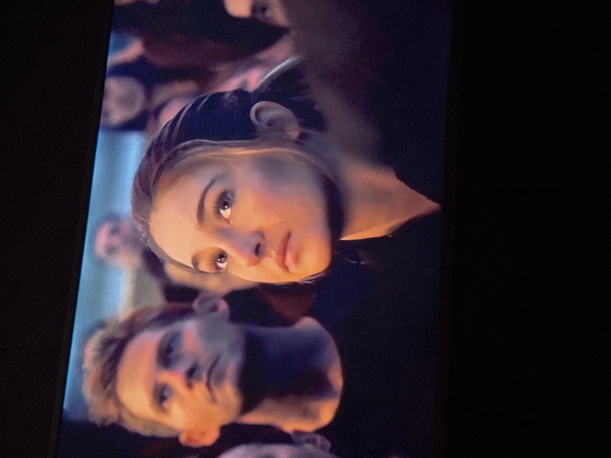 The image shows a close-up shot of a scene with multiple people, focusing on two individuals in the foreground. A fair-skinned woman with light brown hair pulled back, wearing dark clothing, looks upwards with a concerned expression. Her eyes are a light color, and her face is illuminated with a warm light. Below her, a fair-skinned man with short, light hair and a similar dark outfit looks to the left. Both figures are surrounded by a blurred crowd. The environment appears to be dimly lit. The image location is unknown and cannot be inferred from the visual information.