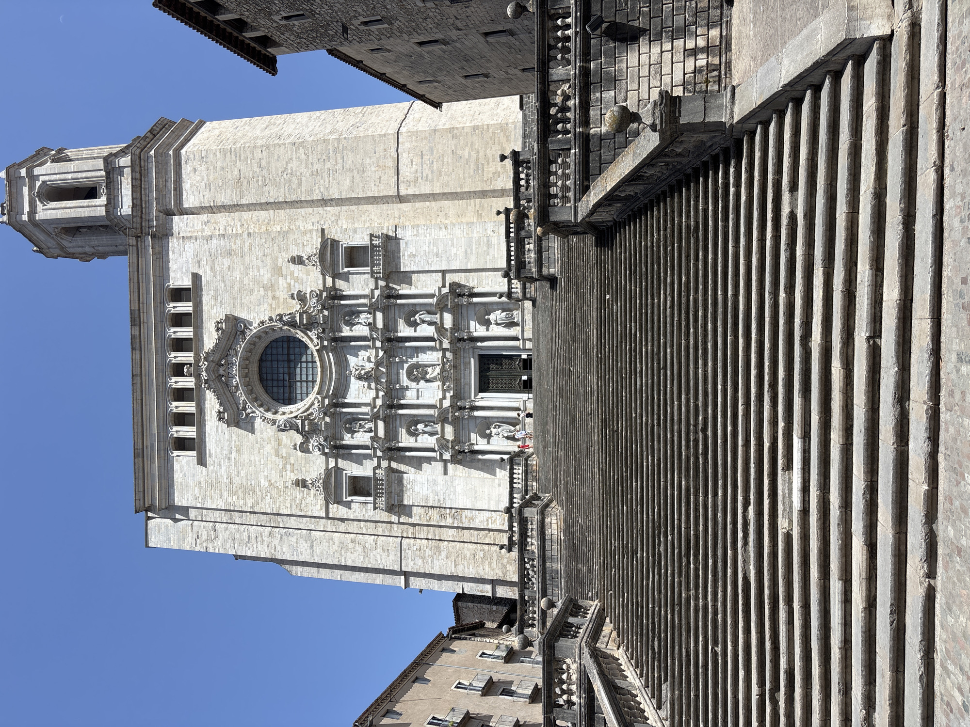 The image captures the imposing facade of a stone cathedral, with a long, grand staircase leading up to its entrance. The cathedral, constructed from pale, textured stone, features intricate architectural details, including a large circular window with ornate carvings, multiple smaller windows, and several statues adorning the facade. The steps of the staircase are wide and made of weathered stone, with a decorative stone balustrade running along one side.
The scene is bathed in bright sunlight, suggesting a clear day, possibly midday given the strong shadows cast. The sky is a vibrant, cloudless blue. In the distance, parts of other buildings with tiled roofs and stone construction are visible, providing context for an urban setting.
There are a few small figures of people visible on the staircase and near the cathedral entrance, giving a sense of scale to the monumental architecture. Their presence suggests that the cathedral is an active place, likely open to visitors. The overall mood of the image is one of grandeur and historical significance. No visible text is present in the image.