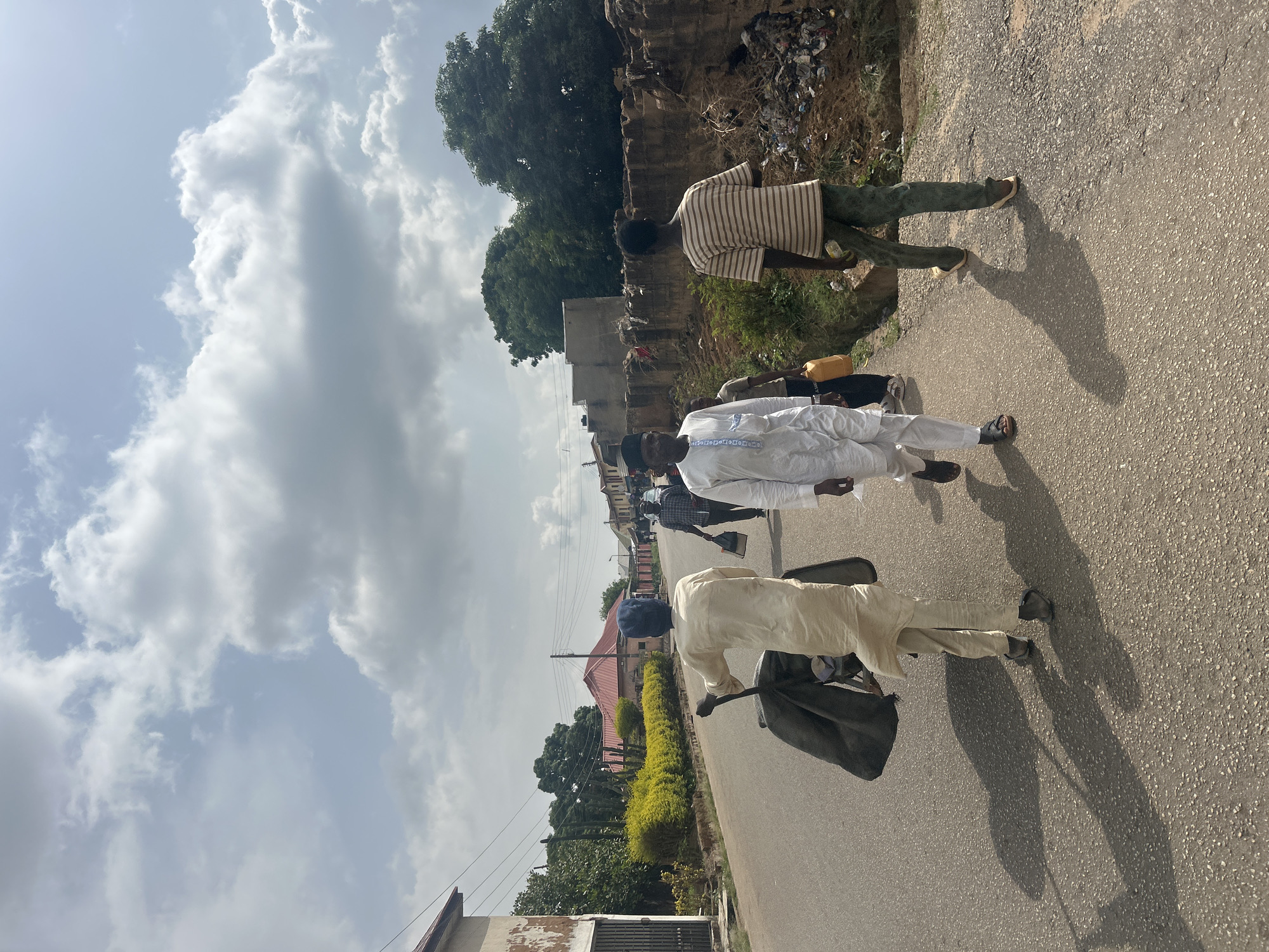 The image depicts a street in Bar Jirgi Summa, Nigeria. Three men are walking along the asphalt road. The first man wears a striped shirt and green pants. The second man is dressed in a white traditional outfit. The third man is wearing a light-colored robe and carries a bag and what appears to be a walking stick. The sky is cloudy. Buildings and vegetation line the side of the road. A small yellow jerrycan is carried by the second man. Debris is visible near a wall along the roadside.