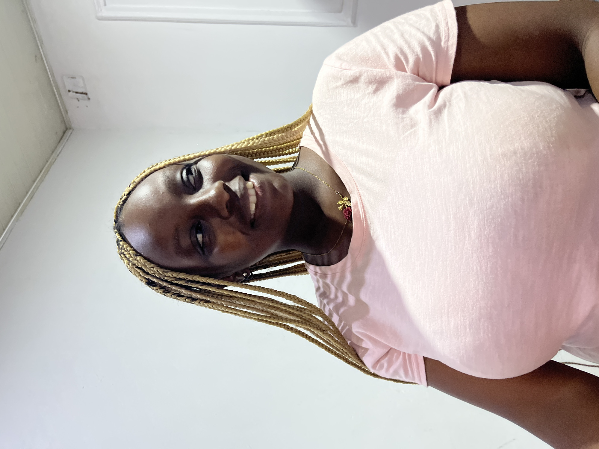 A medium shot shows a dark-skinned woman with braided hair and wearing a pink t-shirt. She is smiling and facing the camera. She is indoors in a location that appears to be a room with white walls. The city and state are Unknown.