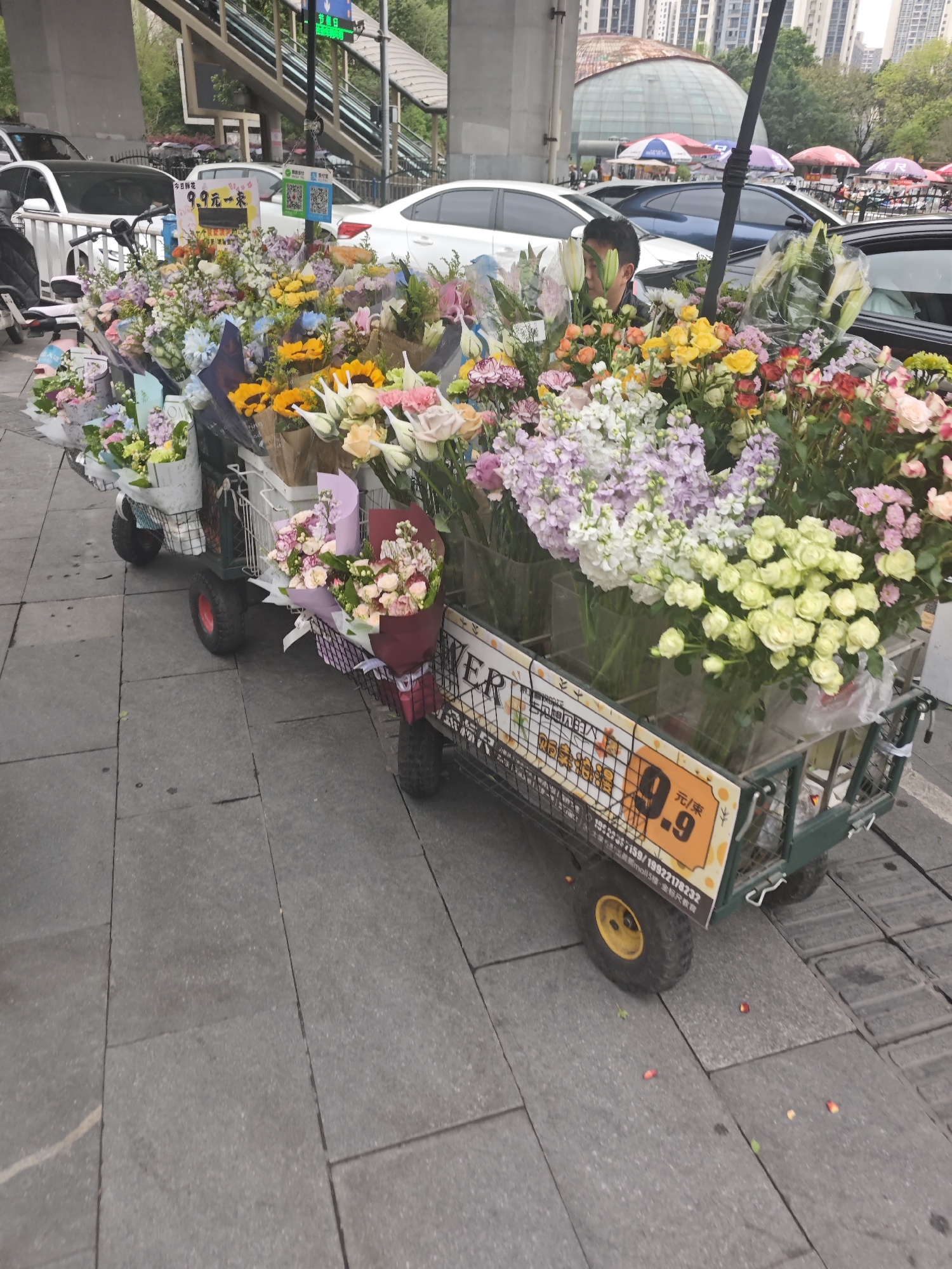 A flower vendor is selling bouquets from a cart in the Chongqing High-tech Zone, China. The cart is loaded with a variety of colorful flowers, including roses, lilies, and sunflowers. A man is partially visible behind the flowers. The cart has a sign advertising flowers for 9.9 yuan per bunch. The scene is set on a sidewalk with vehicles and a pedestrian bridge in the background.