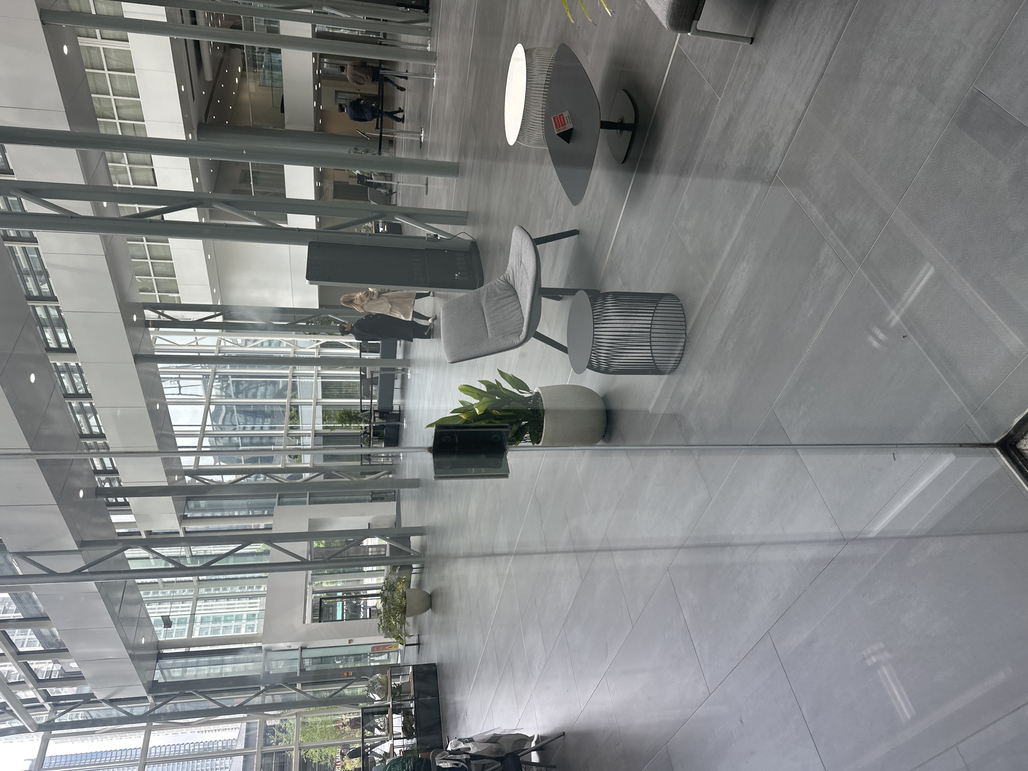 The image shows the interior of a modern building with a glass facade, reflecting an exterior view. The architecture features a structural steel framework. Inside, there is contemporary furniture including a gray chair, side table, and elliptical light. A potted plant is visible. People are walking in the background. The floor is tiled with gray square tiles. The location in the city of Unknown, Unknown is not shown or inferred.