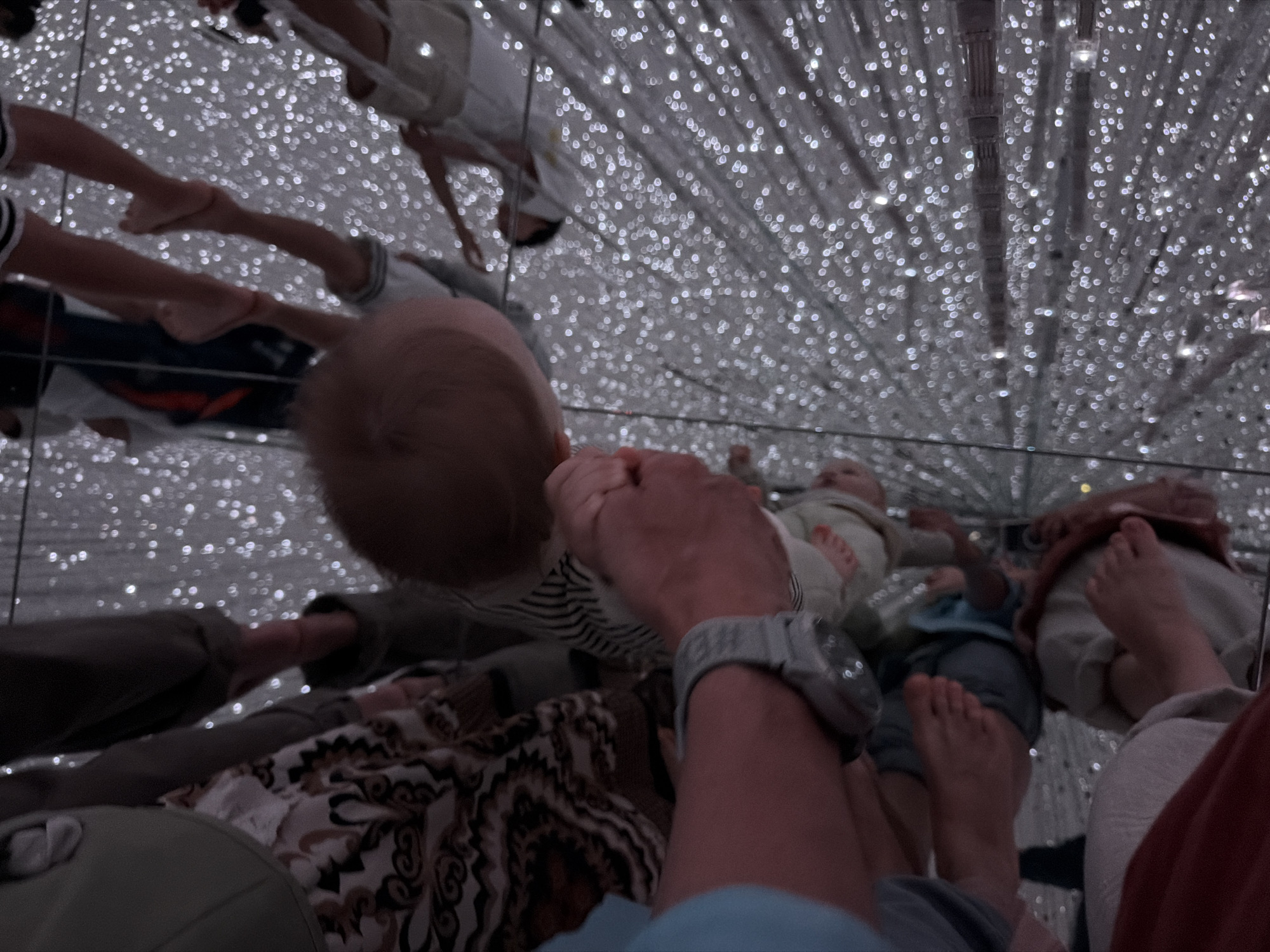 In the city of Koto, Japan, within a digital art museum, a group of individuals are captured in a reflection. The scene is illuminated by numerous small, bright lights that create a sense of depth and wonder. In the foreground, a person's arm, wearing a grey watch, is visible, gently holding a baby. The baby, with its head turned away from the camera, appears to be looking at the surrounding lights. The reflections show multiple pairs of feet, suggesting a crowded environment. Some feet are bare, while others are wearing shoes. The overall impression is one of a family enjoying an immersive and visually stimulating experience in a modern art installation.
