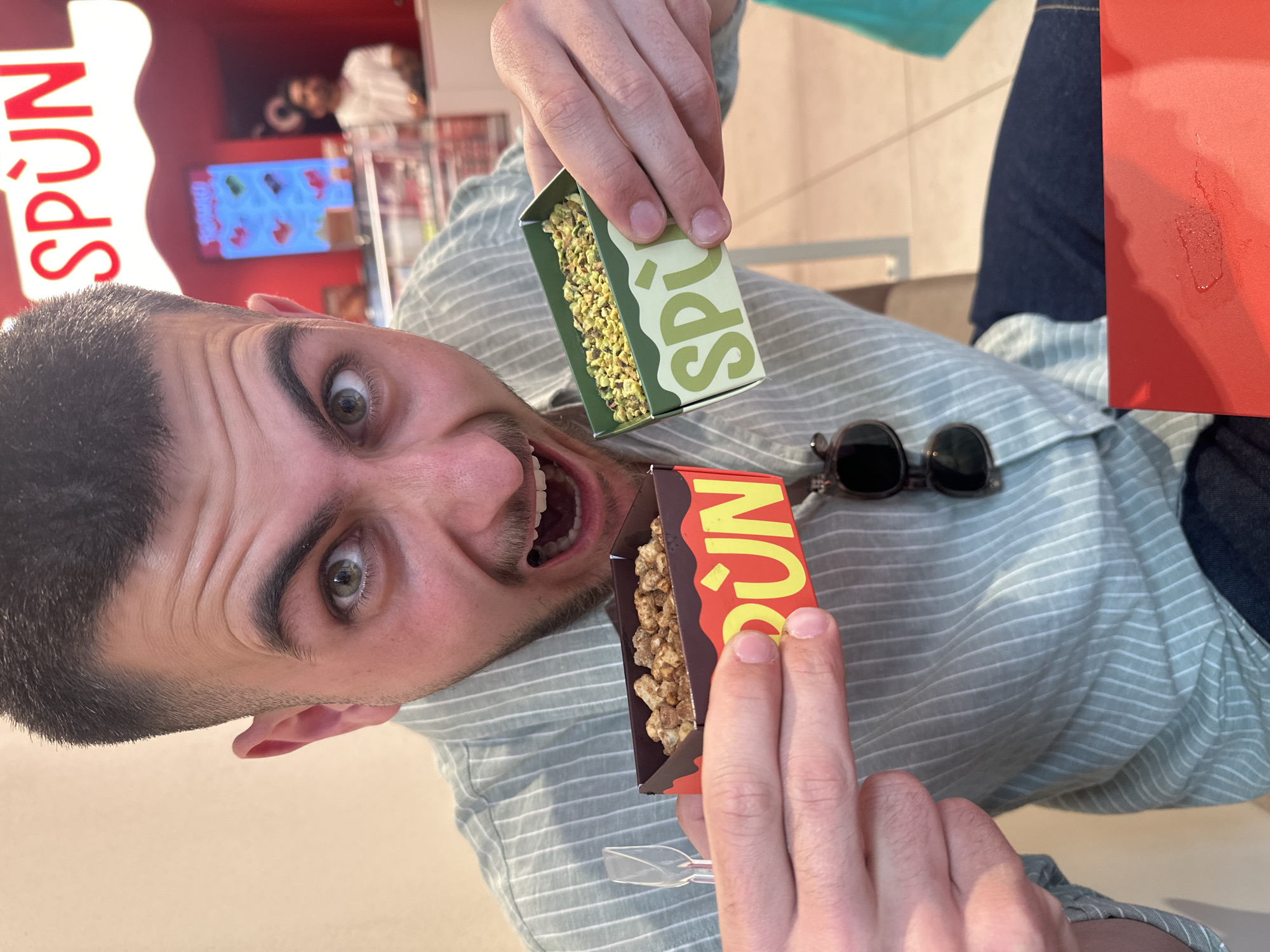 A man is posing with two boxes of Spun popcorn in what appears to be a shop in Milan, Italy.
The man is holding a green box in his left hand and a red box in his right hand, both with the word "SPUN" on them. The boxes contain different flavors of popcorn. He is making a playful, exaggerated expression with wide eyes and an open mouth. He has dark hair, eyebrows, and a beard, and is wearing a light blue striped shirt.
In the background, there is a red structure with the name "SPUN" displayed on it, along with a display screen showing various products. Sunglasses are placed on the shirt.