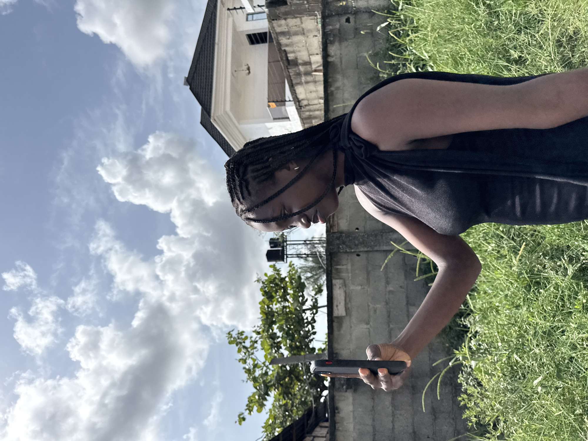 A person with braided hair, wearing a dark top, is holding a mobile phone in their right hand, looking down at it. They are standing outside in a grassy area next to a concrete wall. In the background, there is a two-story building with a white facade and gray roof, as well as some trees. The sky is blue with scattered white clouds. The location is presumed to be Iranla, Nigeria.