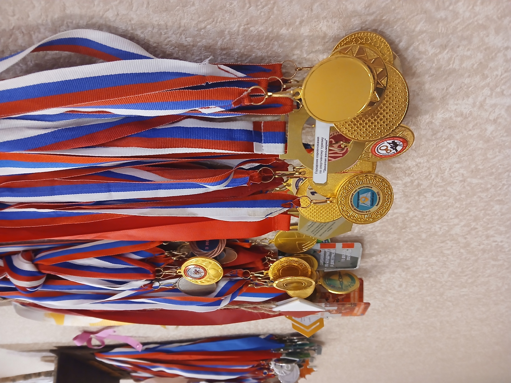 The image shows a collection of gold medals hanging on ribbons with the colors of the Russian flag (white, blue, and red). Some medals have labels indicating they were awarded at competitions for sports tourism called "Golden Eight" in the city of Berezniki. A separate label specifies a competition for bouldering, organized by the "33rd City Tourist Station" taking place on December 13, 2024, in a location called "Tourism House".