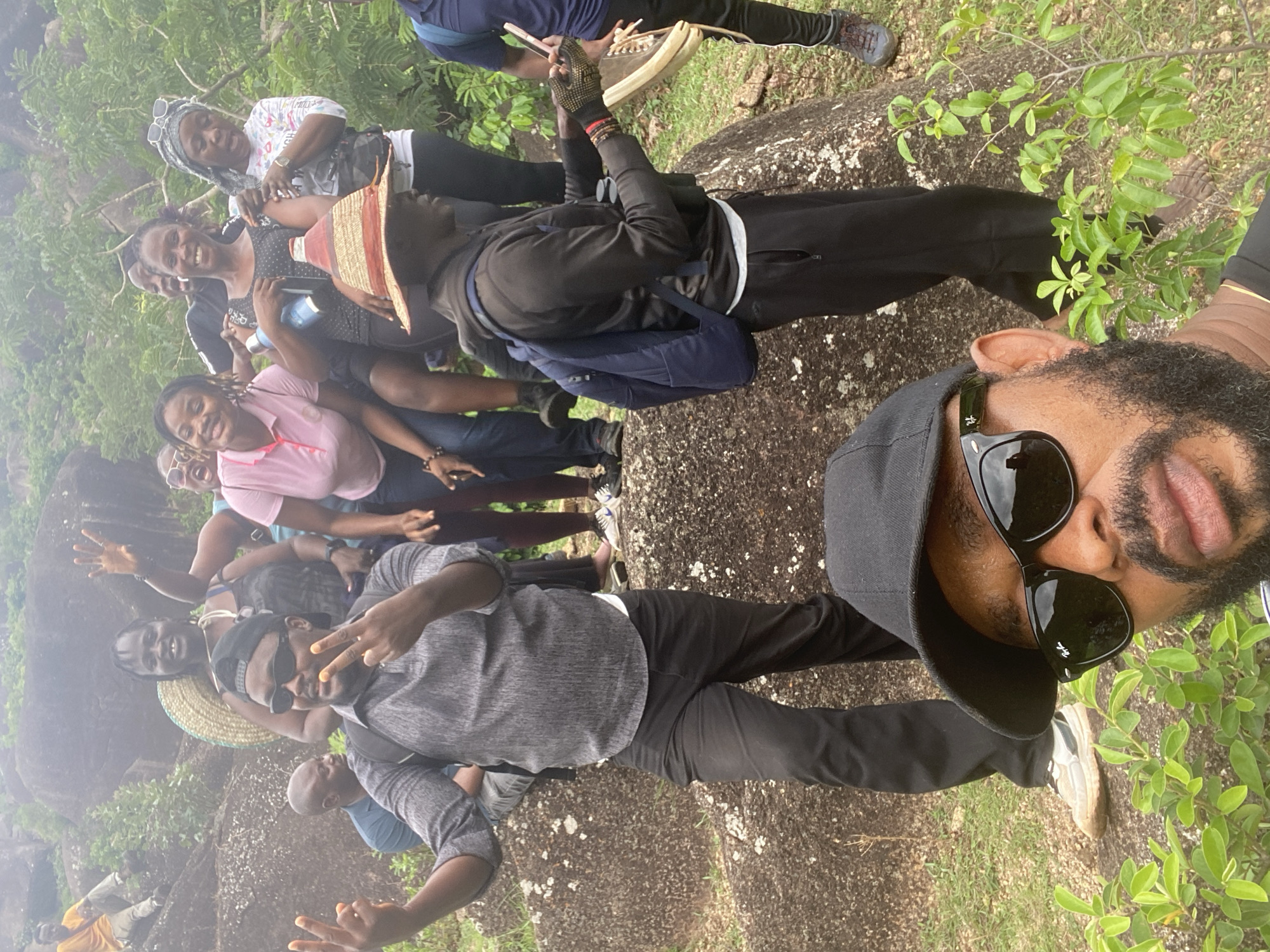 A group of people are posing for a photograph on what appears to be a rock formation, possibly in a rural or natural area near Ganawuri, Nigeria. The group includes men and women of various ages. They are dressed in casual clothing. Some are smiling and making peace signs. A man in the foreground wears sunglasses and a black baseball cap. The environment includes rocks and green vegetation. The background is slightly blurry, possibly due to the depth of field.