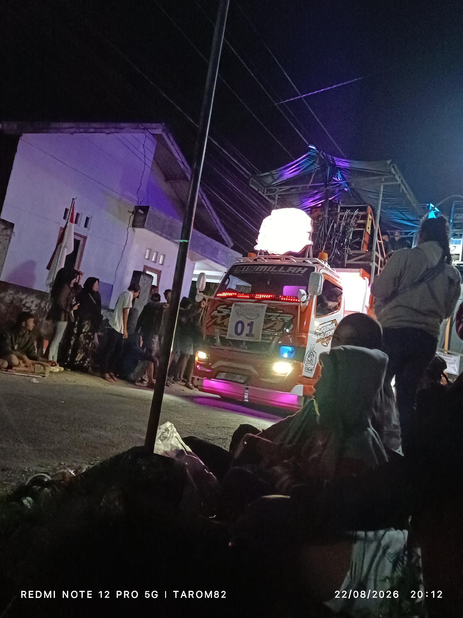At night in Mundusewu, Indonesia, a decorated truck with the number "01" on its side is parked on a street. The truck is adorned with flashing pink and white lights. A large, white inflatable object is mounted on top of the truck, possibly part of a sound system or stage setup. A group of people are gathered on the street, some standing near the truck and others sitting on the ground. The scene appears to be some kind of outdoor event or celebration. A white building with purple lights illuminating its facade is visible in the background. The sky is dark, indicating nighttime. The timestamp on the image indicates it was taken on August 22, 2026, at 20:12.