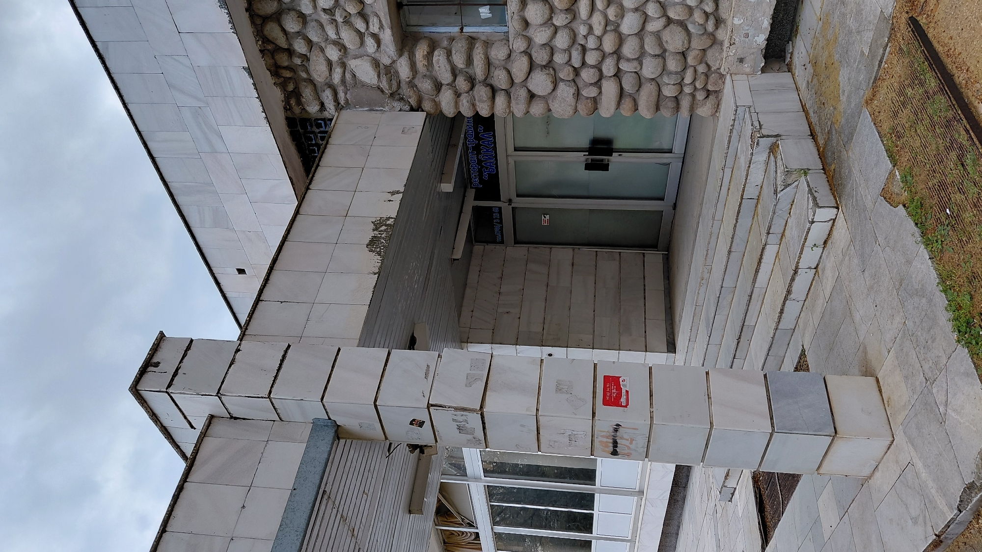 The image depicts a building in an urban environment, with the city and country of its location being unknown. The building's exterior features a combination of materials, including white marble tiles, a textured wall of rounded stones, and glass windows. The building appears to be a restaurant with the sign "БАЙКАЛ" visible above the entrance, which is a set of double glass doors. The entrance is accessible via a short flight of tiled stairs. The surroundings consist of a paved area and a patch of land with grass and a metal fence. The weather appears overcast.