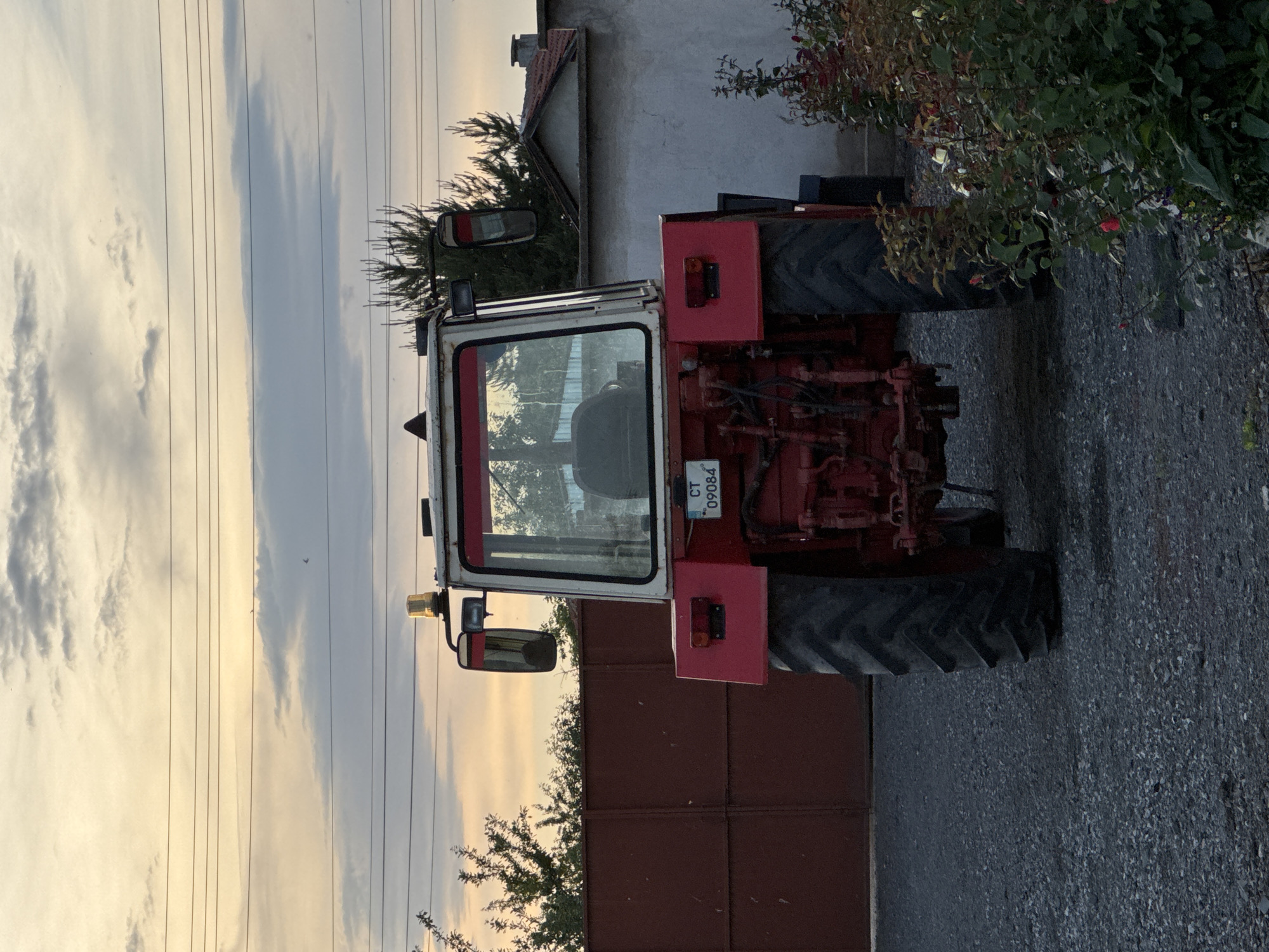 The image shows a red and white tractor parked on a paved surface, possibly asphalt or gravel. The tractor has a cabin with reflective glass. A license plate is visible on the back of the tractor, displaying "CT 09084." Behind the tractor is a building or structure with a white exterior. Adjacent to the building is some greenery, possibly shrubs or bushes. A brown metal fence is also in the background. The sky is overcast with light and dark clouds. The location of the image is the city of Sarnevo, Bulgaria.