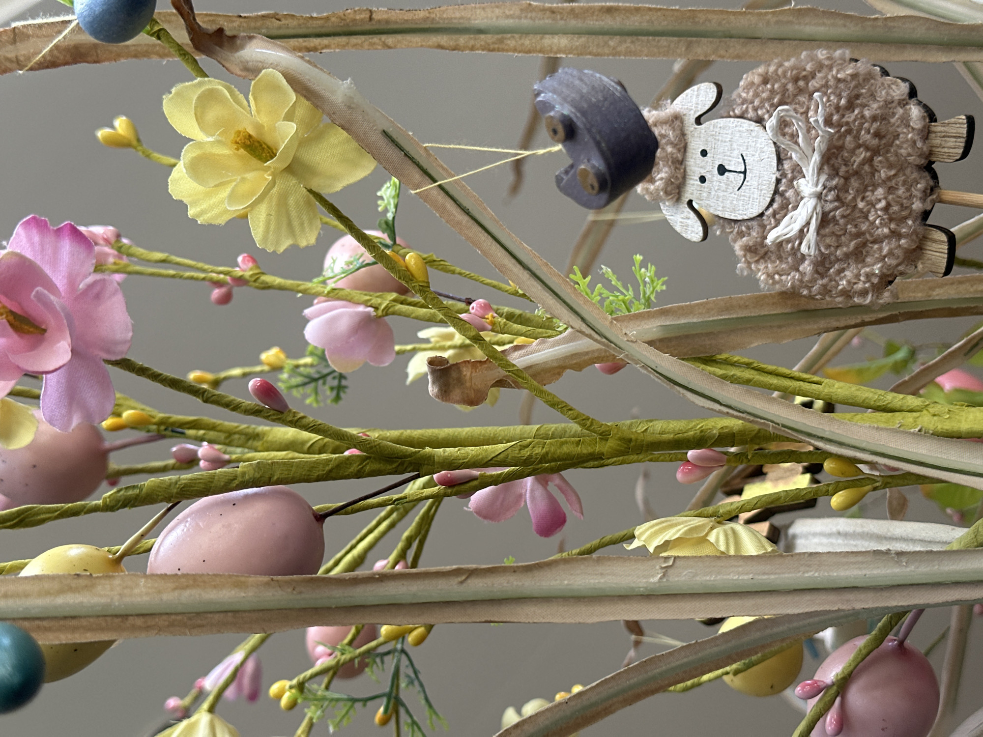 The image shows a close-up of an Easter decoration. It includes pastel-colored Easter eggs, pink and yellow flowers, and a wooden sheep ornament with a smiling face and a tan-colored wool body. The decoration is mounted on faux green branches and light brown horizontal bars. The background is a neutral gray color. The location in Wealden, United Kingdom, is not evident from the image.