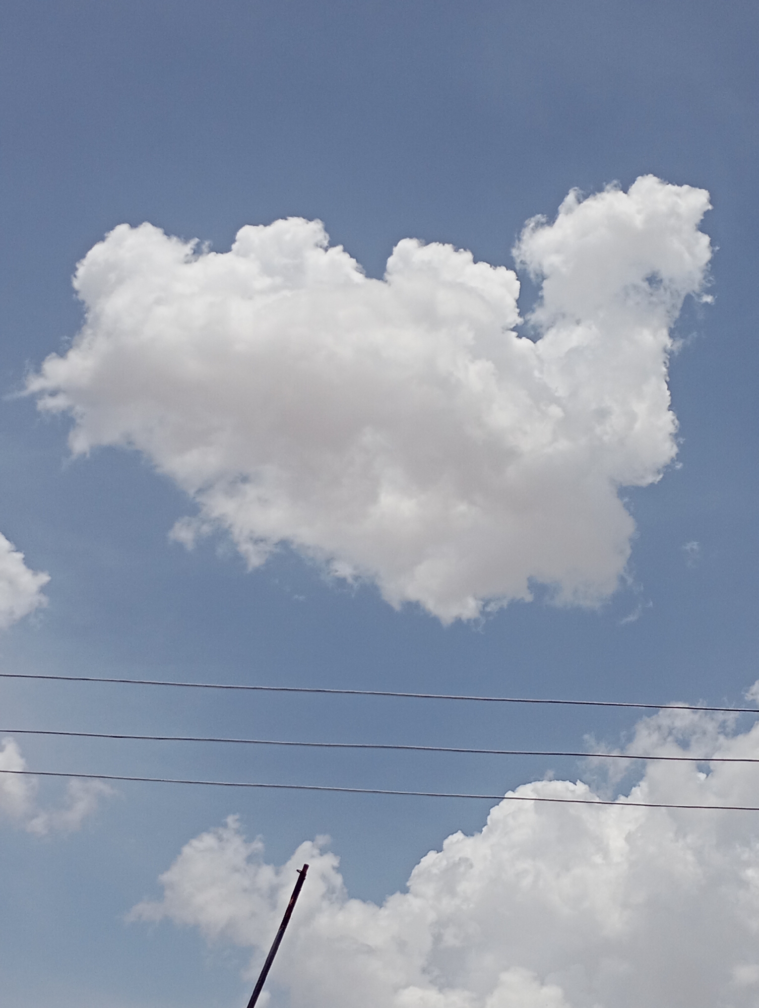The image captures an outdoor scene dominated by a clear, bright blue sky filled with white, fluffy cumulus clouds. A large, prominent cloud occupies the upper-center of the frame, appearing dense and voluminous. Below this main cloud, three dark, parallel power lines stretch horizontally across the mid-ground, extending from left to right. Another, smaller cloud formation is visible in the lower-left, partly obscured by the power lines. In the bottom-right corner, a thin, dark pole or rod is angled upwards into the frame. The scene is brightly lit, indicating a clear, sunny daytime. The overall impression is one of open sky and fair weather. There are no people or discernible activities taking place. The setting is Vom, Nigeria. No visible text is present.