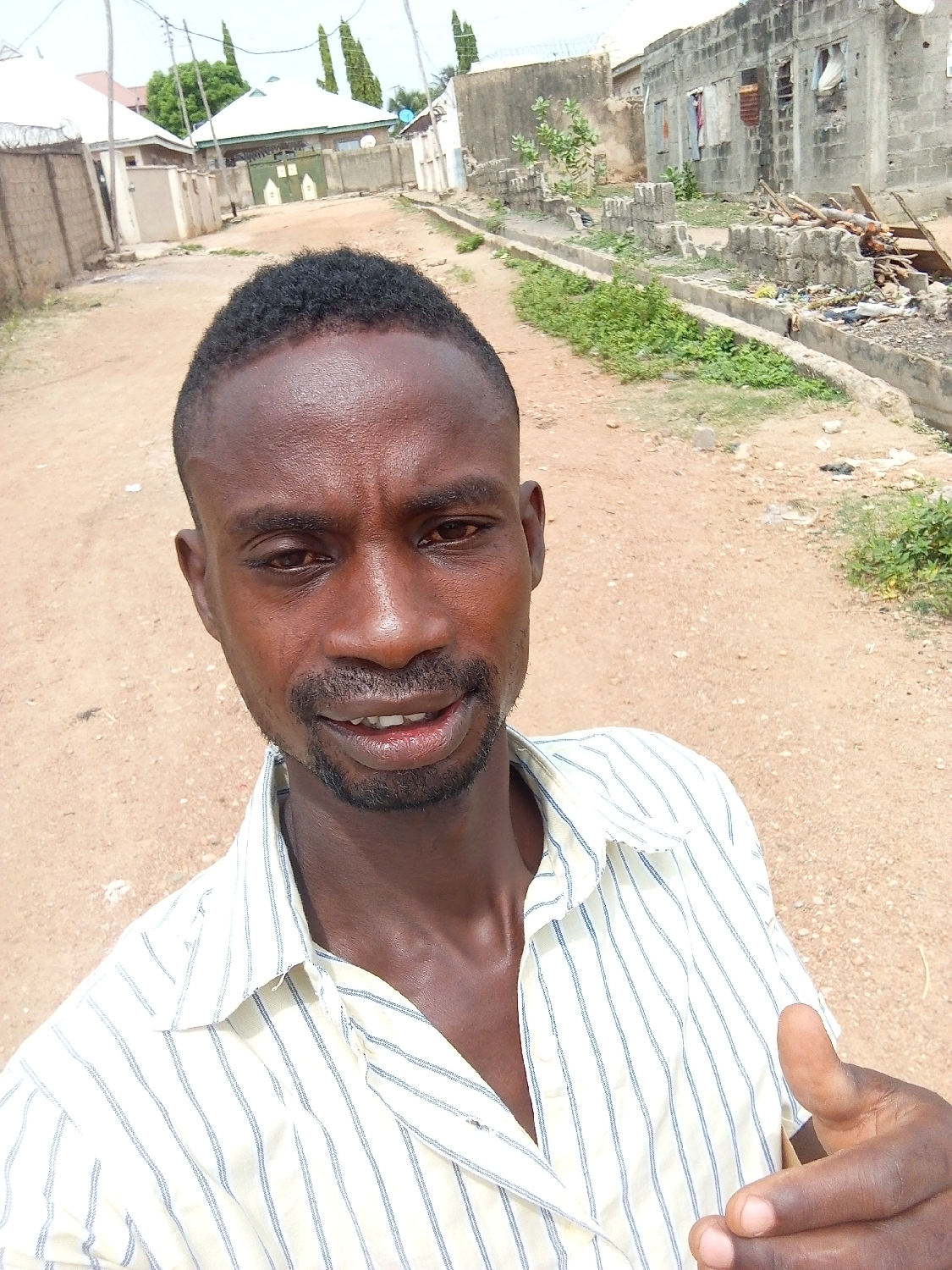 A close-up selfie shows a man of dark complexion looking directly at the camera with a slight smile. He has short, dark, textured hair and a trimmed beard and mustache. He is wearing a white collared shirt with thin vertical blue stripes. His right hand, with a partial thumbs-up gesture, is visible in the bottom right corner of the frame.
The background reveals an unpaved, reddish-brown dirt road stretching into the distance. On the left side, there are fenced properties and several residential buildings with light-colored walls and roofs, one featuring a prominent green-painted gate. Tall, slender trees punctuate the skyline further back. On the right side of the road, the environment appears more rugged, with partially built or dilapidated concrete block structures. A concrete drainage channel runs alongside this section, containing some green vegetation and accumulated debris, including what appears to be construction waste and trash. The sky overhead is bright and clear, indicating it is daytime. The scene depicts a residential street in Adankolo, Nigeria, showcasing a mix of established homes and areas with less development or visible disrepair. There is no visible text.