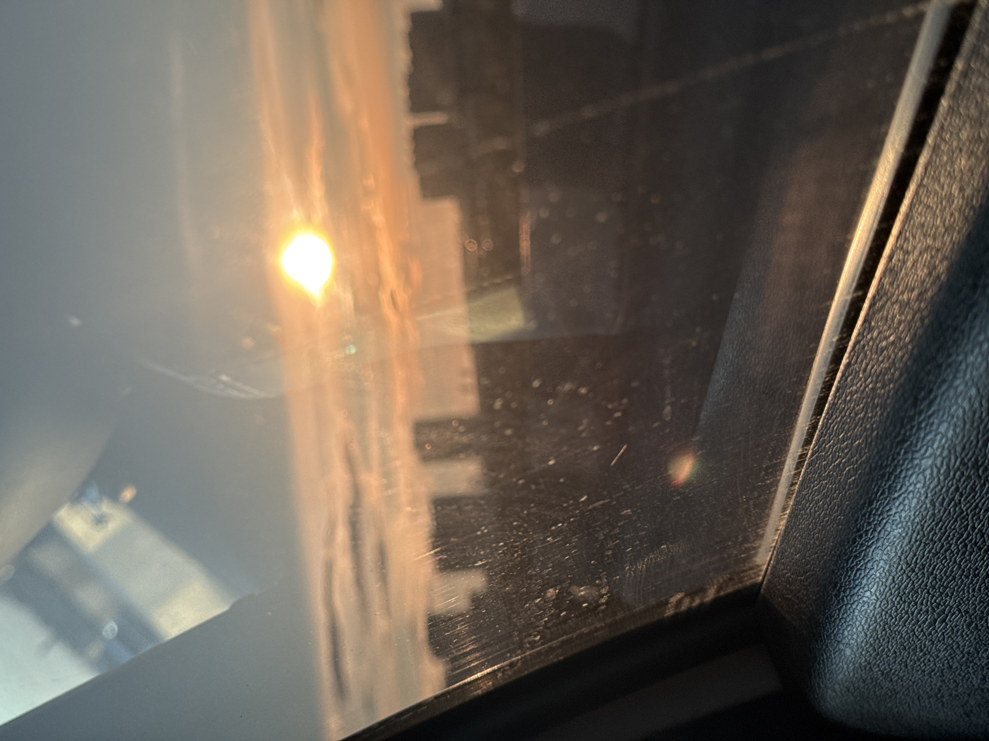 The image shows a view through a car window. The sun is visible, blurred and filtered through the glass. The reflection of buildings or structures is also visible in the window. The interior of the car, including a textured black surface, is visible on the right side of the image. The location in the city is unknown.