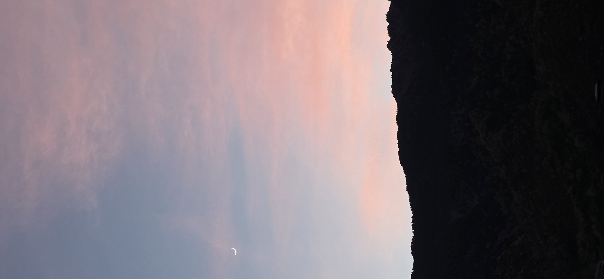 The image captures a twilight scene in Benasque, Spain, featuring a crescent moon in a pastel-colored sky. A dark, silhouetted mountain range occupies the right side of the frame, its peaks and contours distinctly visible against the fading light. The sky transitions from a soft pink near the mountains to a light blue in the lower-left portion, where the moon is situated. The landscape is devoid of artificial lights, suggesting a remote or rural location.
