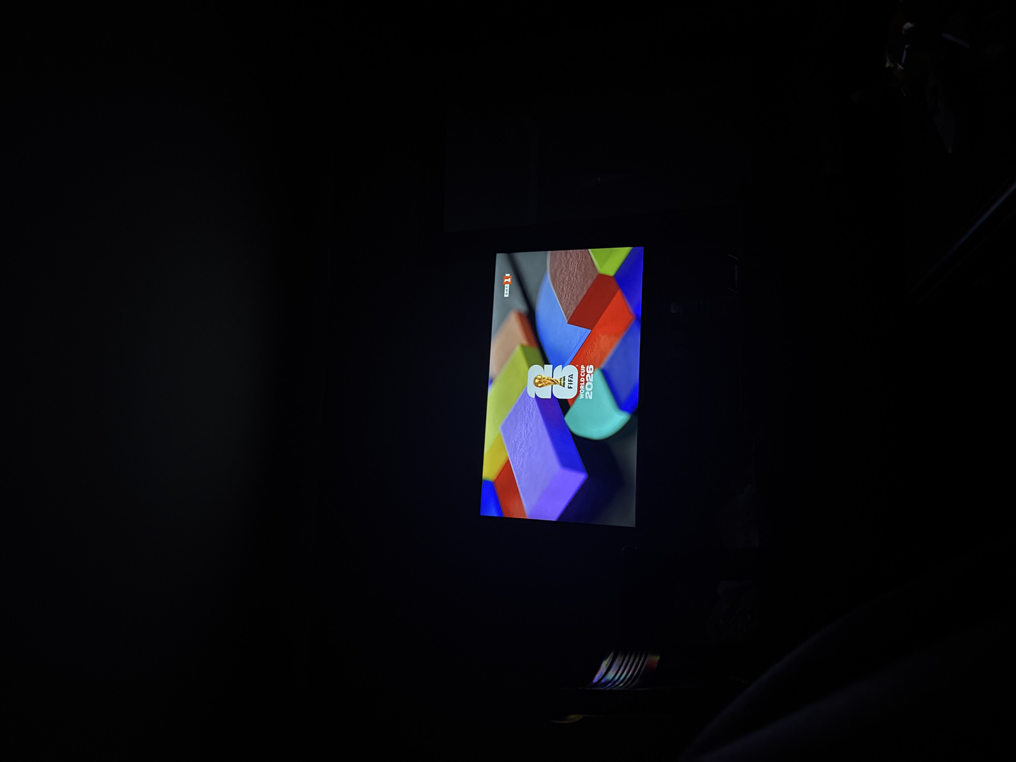 The image shows a television screen displaying a graphic with the FIFA World Cup 2026 logo, along with colorful puzzle pieces. The TV is in a dark room, with some ambient light reflecting off the screen and a portion of a person or object visible in the lower left corner. The scene appears to be indoors, possibly a living room. The location in the city of Belogradchik, Bulgaria is not indicated by any visual elements in the image.