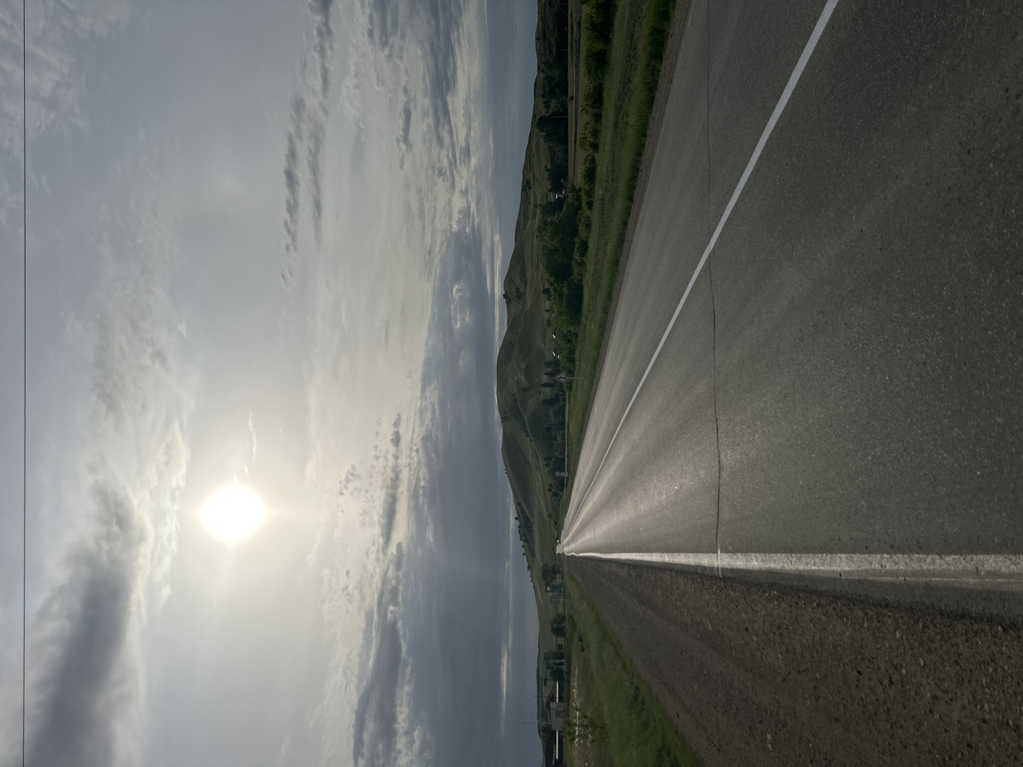 The image presents a view along a straight asphalt road, divided by a solid white line. The road stretches towards a distant hillside, which is covered with green vegetation. To the left of the road, the sky is visible, featuring a bright sun obscured by clouds, with varying shades of gray. A portion of the landscape includes trees, buildings, and grassy areas. The location is indeterminate, but it appears to be in Unknown, Kazakhstan based on the provided context. There is no presence of any people in this image.