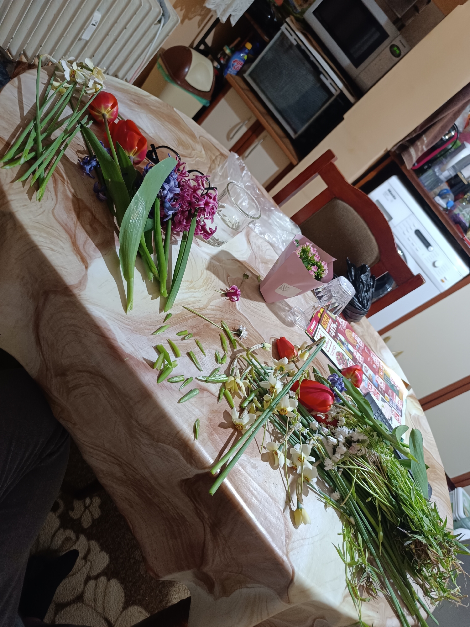 The scene depicts a table covered with various items, including several bunches of flowers, seemingly in the process of being arranged or disassembled. The flowers consist of daffodils, hyacinths, tulips, and possibly other smaller white flowers. Some loose petals and cut stems are scattered on the table.
Additional items on the table include a pink-wrapped small bouquet, a clear vase, a dark plastic bag, a promotional paper or leaflet (potentially a supermarket leaflet or advertisement), and a pair of glasses.
The setting appears to be indoors, possibly a kitchen or dining area, given the presence of kitchen appliances such as a microwave and a washing machine visible in the background. There is also a chair with a brown fabric seat partially visible.
Based on the information provided, the location is Pleven, Bulgaria. However, the image itself provides no specific clues confirming this location. The description is based solely on the visible elements within the image.