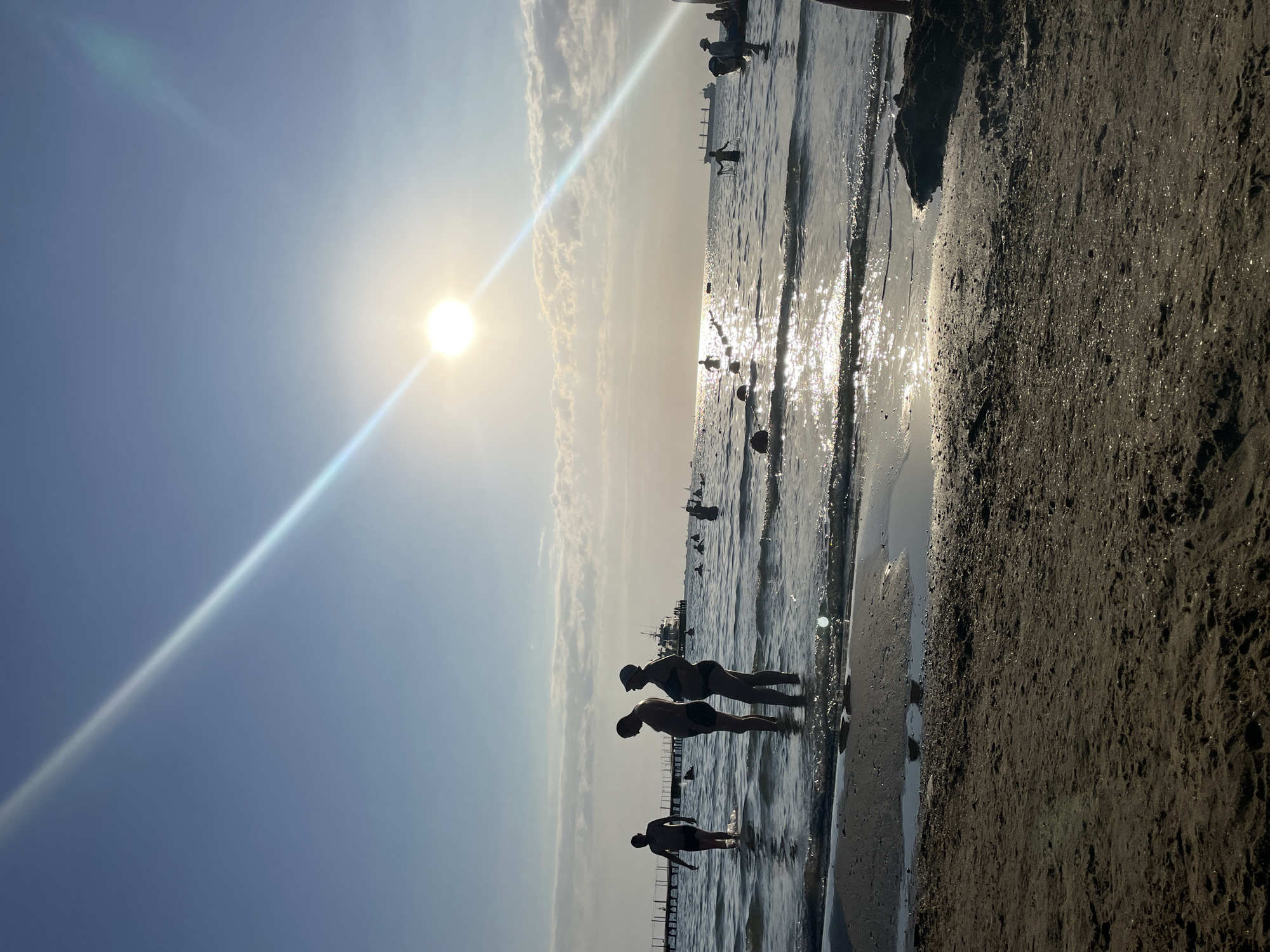 The photograph, taken at a beach in Anapa, Russia, captures a serene late afternoon scene by the Black Sea. The sun, low in the sky, casts a bright glare and a prominent light streak across the frame, illuminating the water's surface with a shimmering reflection. Several people are visible in the shallow water near the shore. In the foreground, two individuals stand facing the sea, their bodies silhouetted against the bright light. Further to the left, another person walks in the water. In the middle ground and background, numerous other figures are scattered throughout the sea, some appearing to swim or wade. A wooden pier extends into the water on the left side of the image, with a boat and what appears to be a large cargo ship visible in the distance. The water is calm with gentle waves lapping at the sandy shore, which occupies the bottom portion of the frame. The sky is partly cloudy, with soft white clouds drifting above the horizon.