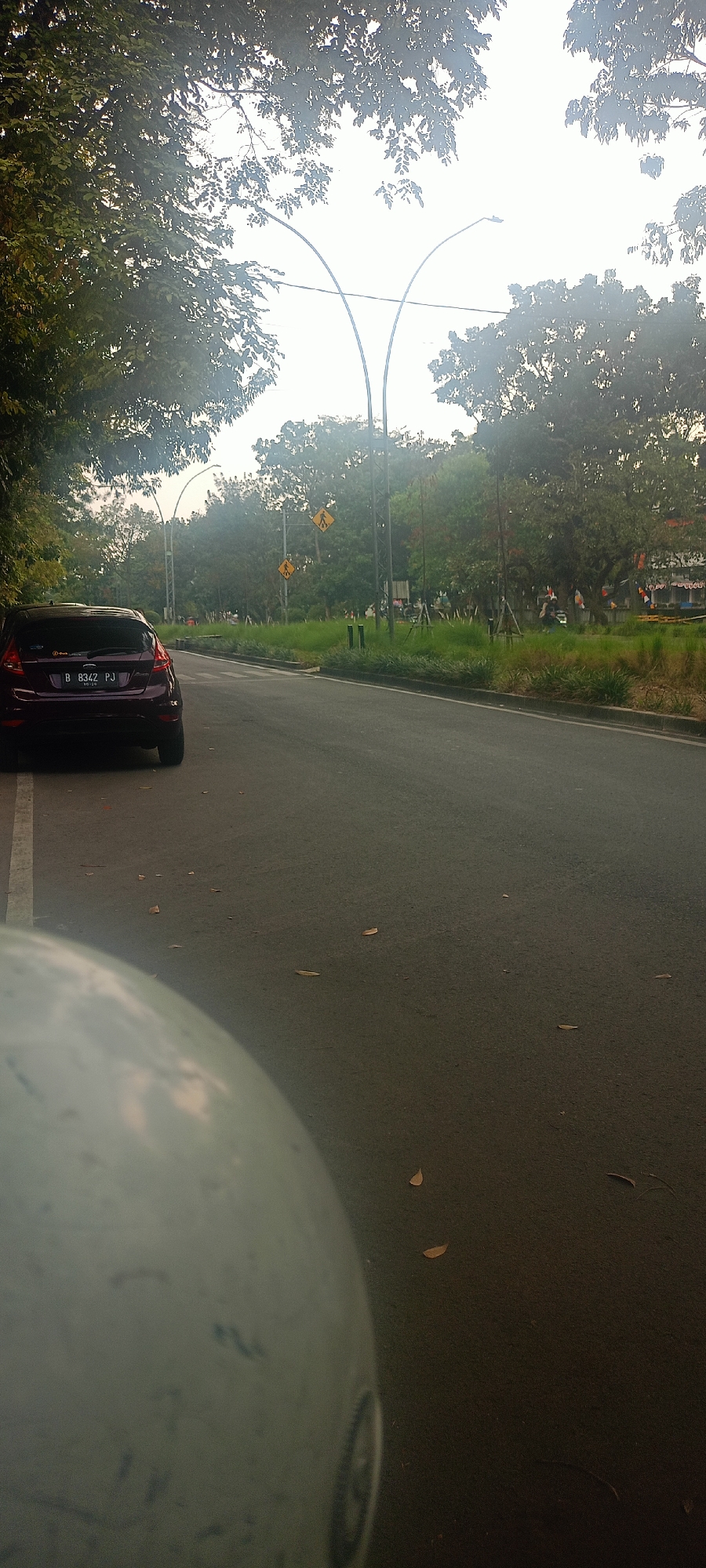 The image shows a street scene in Bandung City, Indonesia. In the foreground, a portion of a light blue helmet is visible, suggesting the perspective of a motorcyclist. The road is asphalt, with a white line marking the edge of the lane on the left. A dark purple car, a Ford Fiesta with license plate B 8342 PJ, is parked on the left side of the road. Beyond the car, the road curves to the right.
On the right side of the road, a grassy area is present, with several trees and some decorative fencing. Two yellow pedestrian crossing signs are visible in the distance. The sky is overcast and bright, indicating daylight. In the background, there are more trees and some buildings, though they are not clearly discernible. The overall impression is that of a suburban or park-like area within the city.