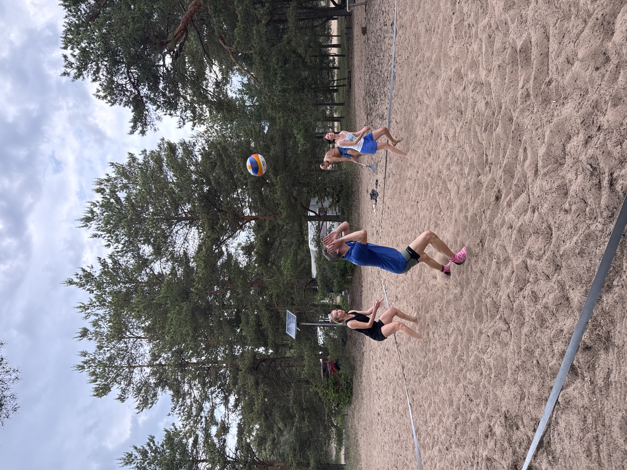 The image displays a beach volleyball court situated in a sandy clearing within a pine forest, under a cloudy sky, in the city of Zelenaya Roshcha, Russia.
Four individuals are present on and around the sandy court. In the left foreground, a woman with blonde hair, wearing a black tank top and black shorts, stands barefoot in a ready position with knees bent and arms slightly forward. To her right, in the center foreground, a man with dark hair, wearing a blue short-sleeved shirt, grey shorts, and pink athletic socks with dark shoes, is in the process of setting a multi-colored FIVB volleyball, with his hands cupped above his head as the ball hovers above him.
Further back on the right side of the court, a second woman with dark hair, wearing a white tank top and blue shorts, stands barefoot in a ready stance, looking upwards towards the volleyball. Behind her and slightly to the left, a shirtless man with short hair, wearing blue shorts, holds a metal rake in his right hand, observing the activity on the court.
The court itself is marked by flat, dark-colored boundary lines visible in the foreground and extending into the middle ground. The ground is composed of light brown, rippled sand with visible footprints.
The background is dominated by numerous tall pine trees with dark green needles and reddish-brown trunks, forming a dense forest. Through the trees on the left, a light-colored recreational vehicle or camper trailer is partially visible. Further left and behind the court, a metal pole with a solar panel attached is installed in the sand. A red vehicle is partially obscured by foliage further back on the left. On the far right, behind the court, a wooden structure, possibly a bench or table, is faintly visible, along with some dark objects (possibly shoes) and the rake previously mentioned, resting on the sand. The sky above is overcast with a mix of grey and white clouds.