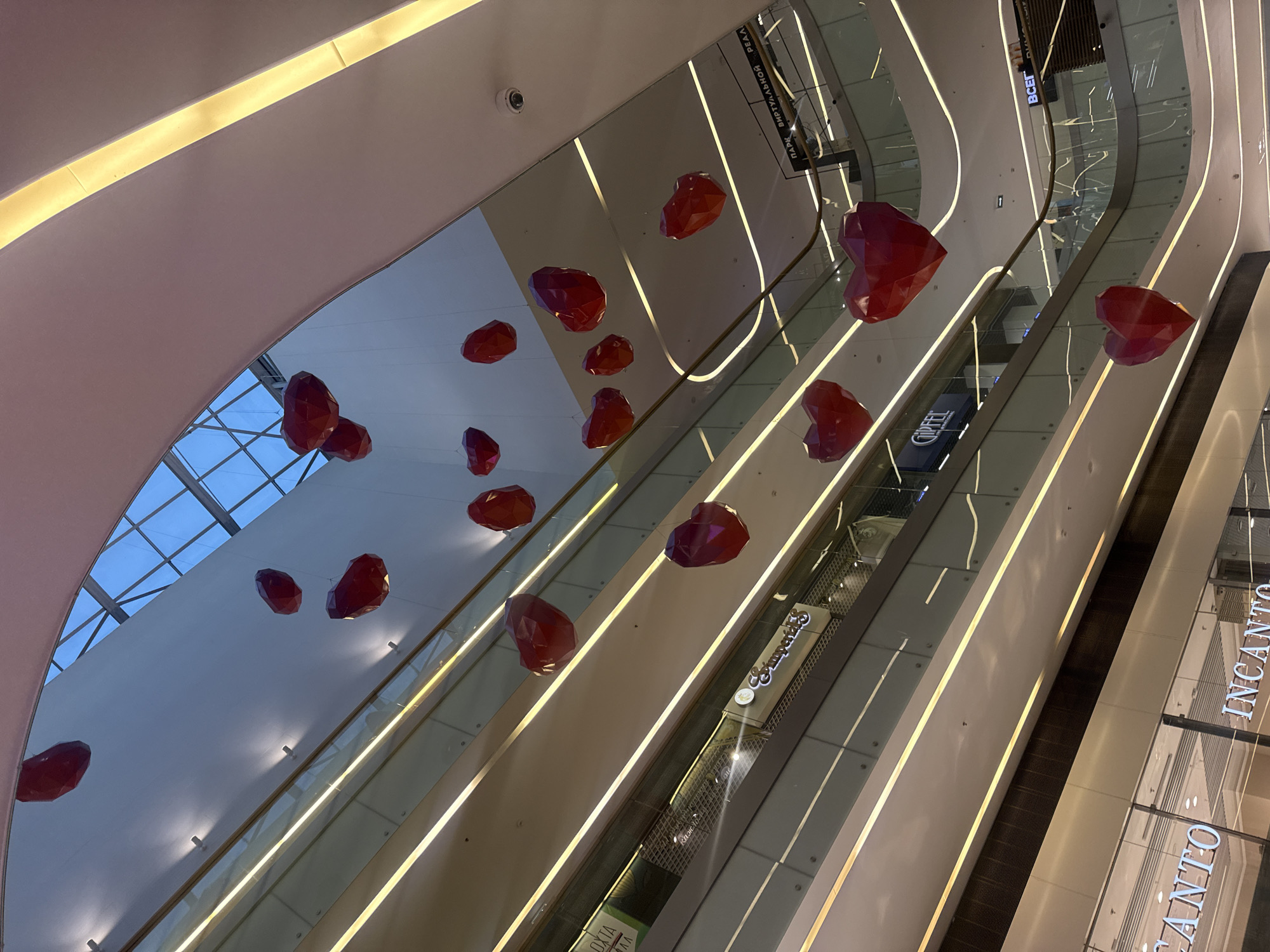 This is an interior shot of a modern shopping mall in Saint Petersburg, Russia. The image looks upward, showcasing the multi-level design of the mall. Red, geometrically-shaped hearts are suspended from the ceiling as decorations. Escalators and glass railings define the different levels. Store signs such as "Incanto" and "COFFEE" are visible. Artificial lighting is integrated into the architectural design with linear lights accentuating the structure. A glass ceiling section allows natural light to enter the space. There are no people visible in the scene.