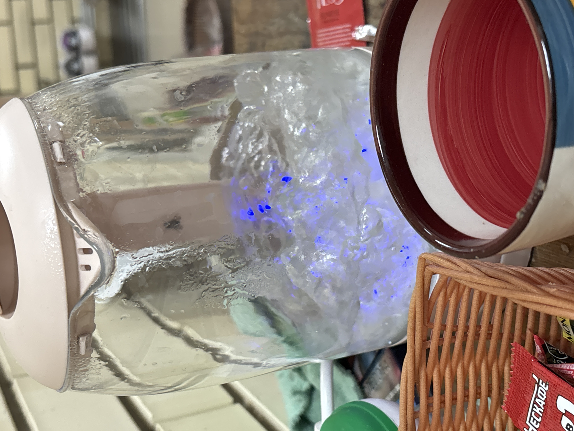 A clear glass electric kettle with a beige-pink top is shown mid-boil, its water illuminated by a vivid internal blue light and visibly turbulent with numerous bubbles. Condensation droplets adhere to the inner glass surface above the waterline. In the immediate foreground, a light-brown woven basket contains a red sachet with partial labeling, including "HECKADE." Adjacent to the basket, a ceramic mug reveals a white interior with a distinctive red, concentric-ring pattern at its base, and a dark brown rim. A light green textile is visible behind the kettle. The background features a light-colored brick wall, with blurred objects, possibly shoes, on a shelf to the far right. No individuals are present in the scene.