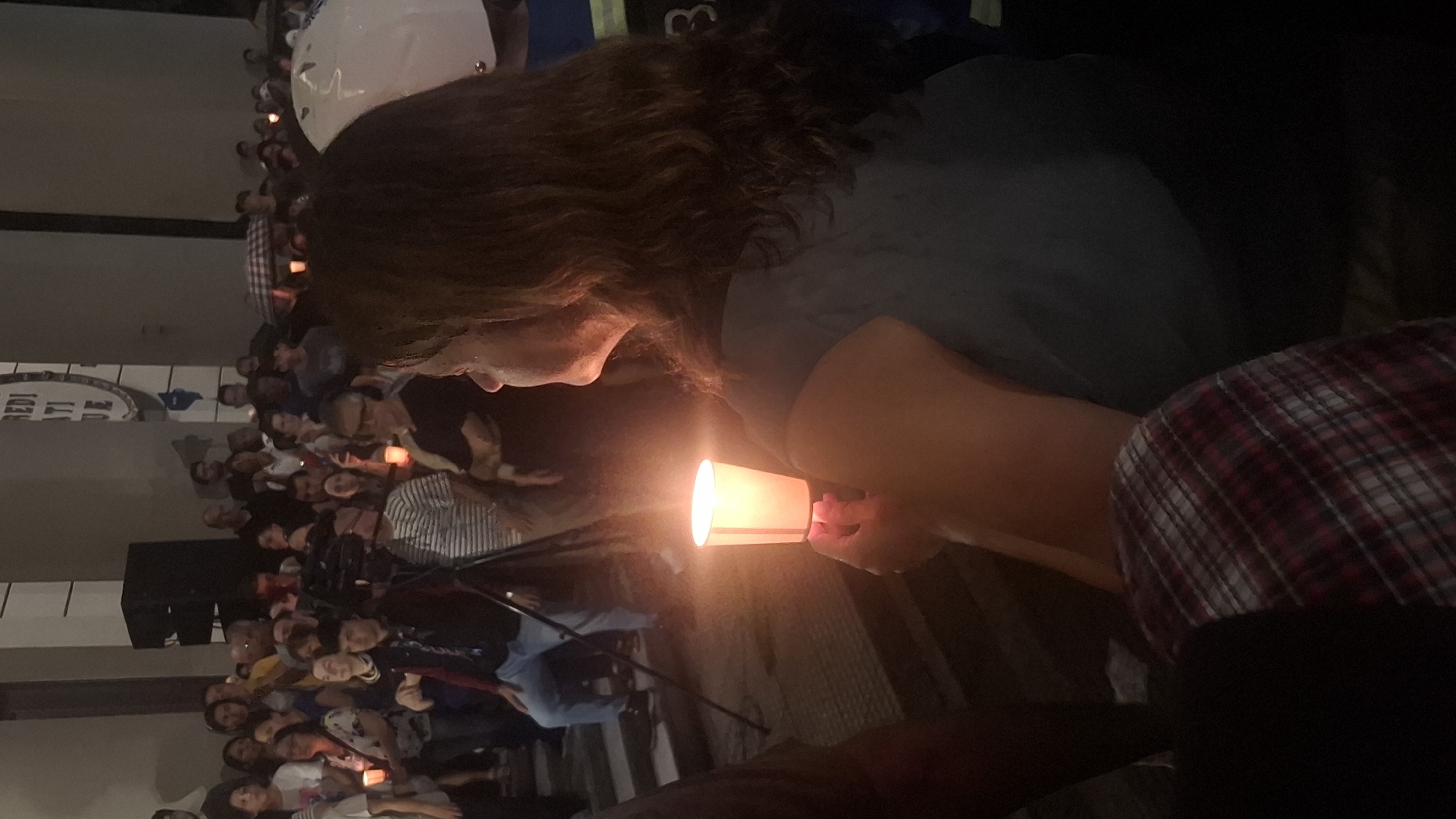 A nighttime image captures a woman holding a lit candle in a paper cup, surrounded by a crowd of people, many of whom also hold candles. The scene appears to be a vigil or memorial. The location is suggested to be Buenos Aires, Argentina, based on the environment.