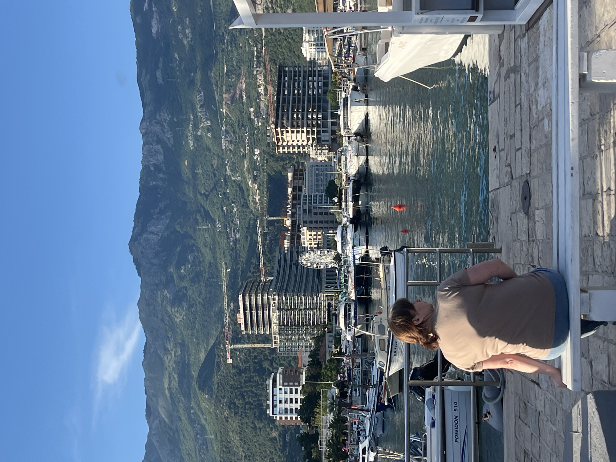 The image captures a panoramic view of a coastal town, likely Budva, Montenegro, with a person seated in the foreground observing the scenery. The photo is rotated 90 degrees clockwise, showing a clear blue sky on the left and a large, green, rocky mountain range dominating the background.
In the immediate foreground, a person with light brown hair, wearing a beige t-shirt and blue jeans, sits with their back to the camera on a white bench or railing, looking towards the water. To their left is a paved stone walkway or dock.
The midground features a bustling marina filled with numerous boats of various sizes, including motorboats and yachts, docked along the waterfront. The water reflects the sunlight, creating shimmering patterns. Visible on one boat is the text "POSEIDON 510" and on another, partially visible, "BD 8322," likely a registration number. Red buoys float in the water.
Beyond the marina, a mix of modern and older buildings line the coast and ascend the lower slopes of the mountains. Several multi-story buildings, some appearing to be under construction with exposed frameworks, and prominent construction cranes indicate ongoing development.
The time of day appears to be late afternoon or early morning, characterized by bright sunlight and clear skies. The overall atmosphere is peaceful, suggesting a moment of relaxation by the water in a scenic, mountainous coastal setting.
