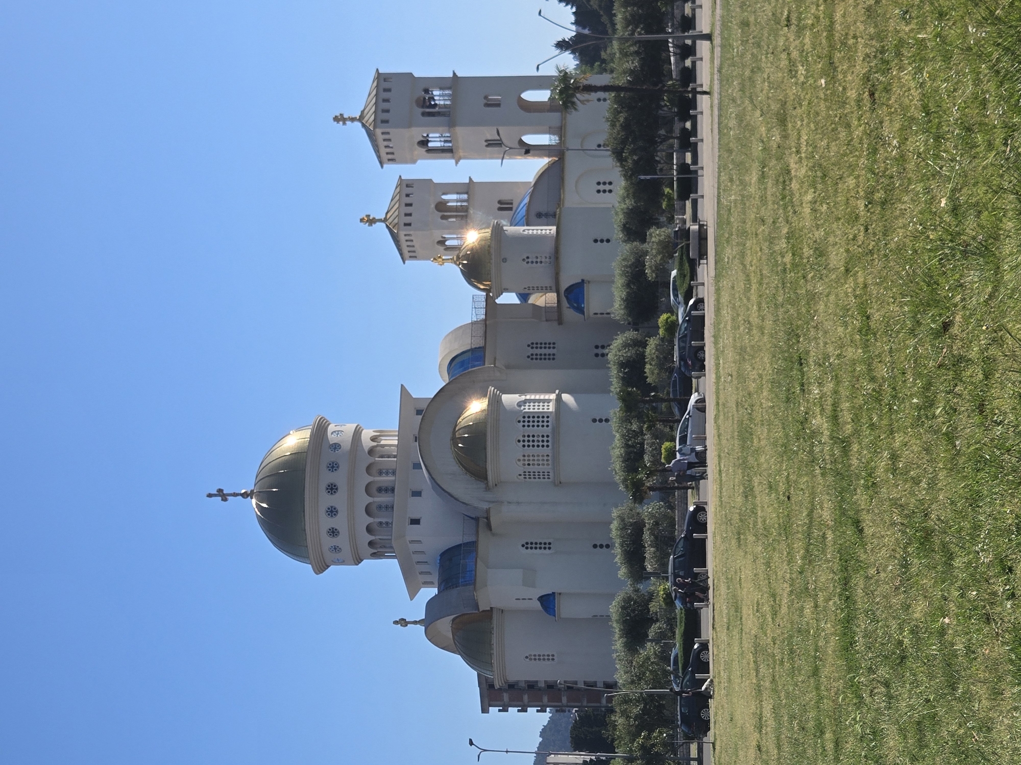 The image shows a church in Bar, Montenegro, with a tall, white structure featuring multiple domes with golden tops. The church has towers with arched openings. The scene includes a grassy area and a street with parked cars and trees. The sky is clear and blue.