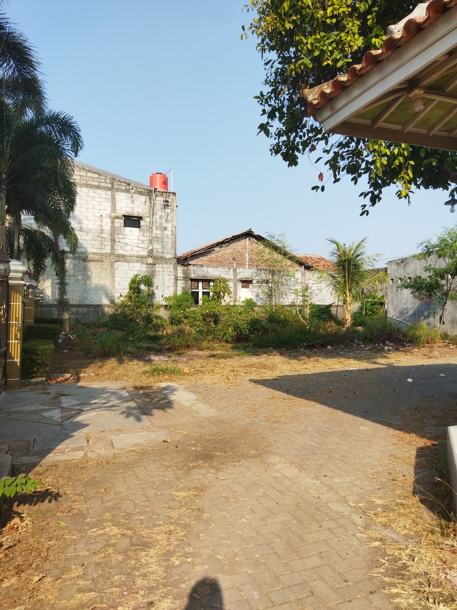 The image shows an outdoor scene with several buildings and vegetation. In the foreground, there is a paved area with interlocking bricks and some dry grass. A shadow of a person is visible on the ground in the lower right corner. To the left, there is a paved walkway with a patterned design.

In the middle ground, there is a patch of overgrown vegetation and small trees. Behind the vegetation, there are several buildings. One building, on the left, appears to be constructed from white concrete blocks and has a red water tank on its roof. Another building, in the center, has a brick roof and visible windows.

In the background, the sky is clear and blue. Trees with green leaves are visible, some of them casting shadows on the ground. The overall impression is of a somewhat unkempt outdoor space in a residential or semi-commercial area.