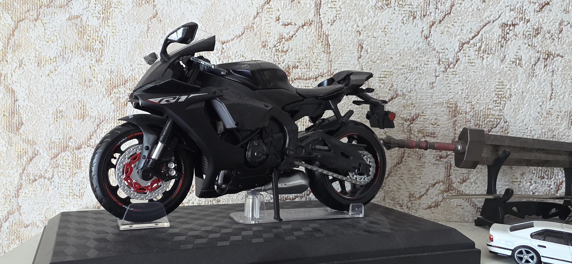 The image depicts a collection of models displayed on a surface against a textured wallpapered background. The primary focus is a black motorcycle, specifically a Yamaha R1 model, with red accents on its wheels. It is mounted on a clear plastic stand. To the right of the motorcycle, a large, dark-colored sword with a red-banded hilt rests on a black stand. Further to the right, a white car model, resembling a BMW, is partially visible. The scene appears to be an indoor display, likely a shelf or table. There is no indication of the location being in Sumgayit, Azerbaijan, or any specific city.