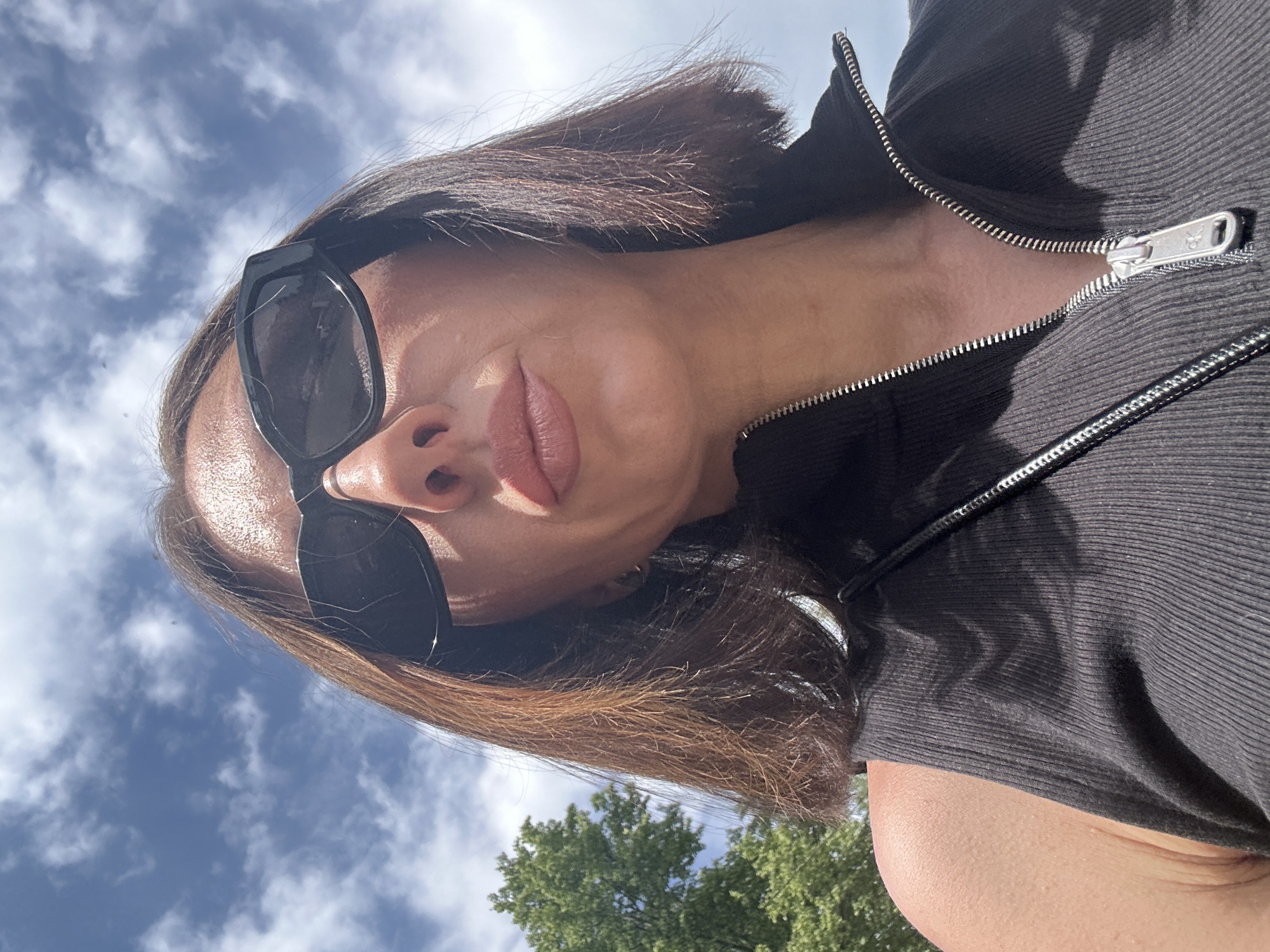 A woman wearing sunglasses and a black zippered shirt is looking at the camera from a low angle. Behind her are trees and a blue sky with scattered clouds. There is no information available to determine if this image was taken in Votkinsk, Russia.