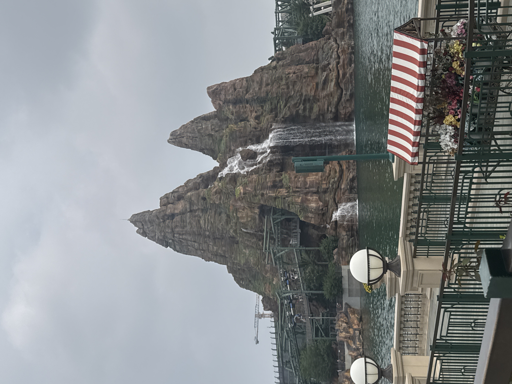 The image depicts a theme park attraction with a large, man-made rocky mountain featuring waterfalls. A roller coaster track snakes around and through the mountain, with several roller coaster cars visible on the track. The attraction is situated on a body of water, with a decorative railing and lampposts in the foreground. A small cart with a red and white striped awning, filled with flowers, is positioned near the railing. The sky is overcast. The scene is located in the Universal Studios Beijing, which is in Tongzhou District, China. No people are clearly identifiable in the image.