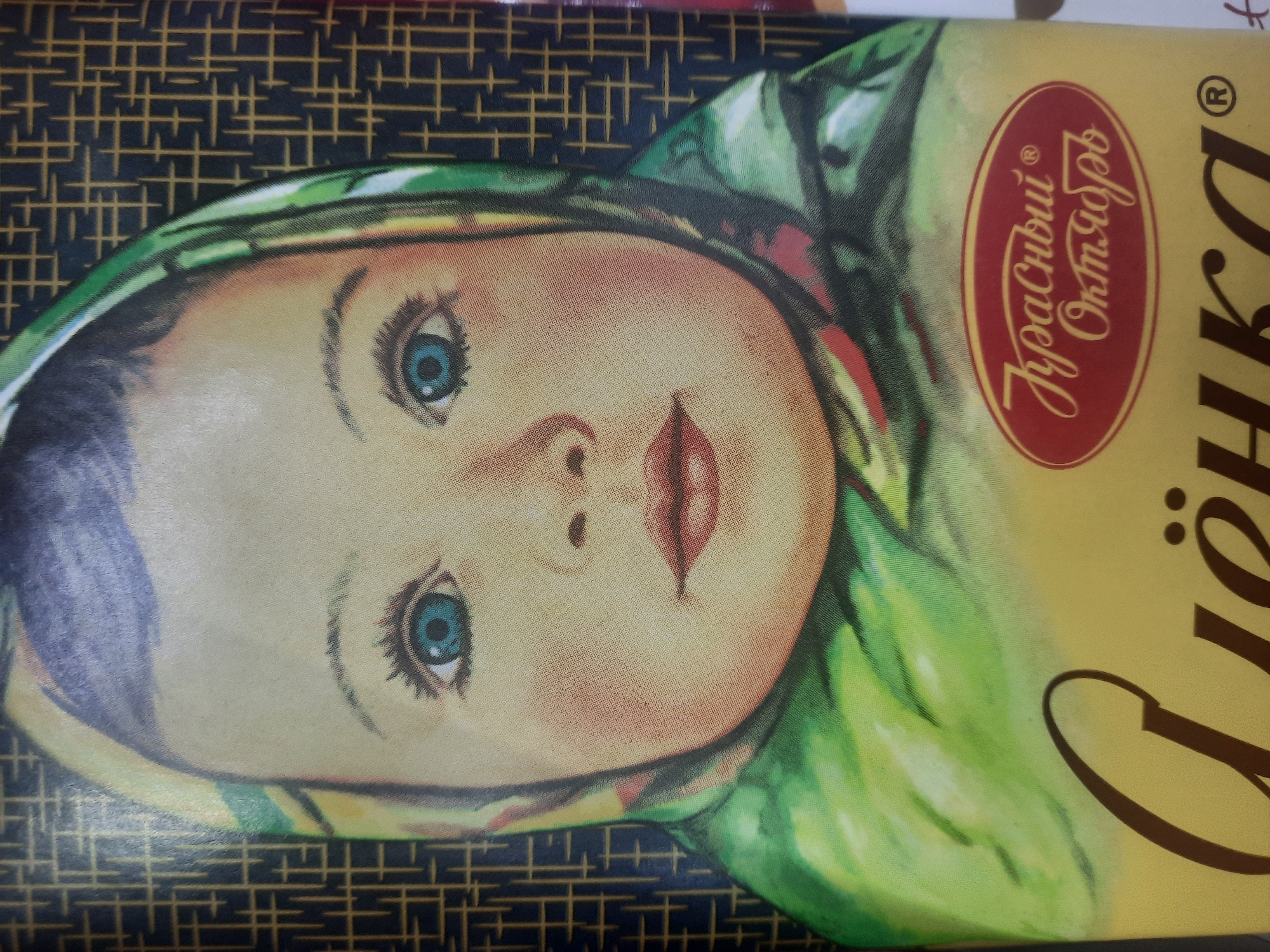 The image shows a close-up of a package featuring a stylized illustration of a baby's face wearing a green and yellow headscarf. The baby has bright blue eyes, rosy cheeks, and pink lips. The background of the package is a dark, patterned material. Below the illustration, a logo in a red oval contains Cyrillic text that reads "Красный Октябрь" (Krasny Oktyabr). Below the logo, the word "Алёнка" (Alyonka) is written in large, brown, cursive letters, with a registered trademark symbol to the right. The overall impression is of a confectionary product packaging. The location in the city of Парэчча, Belarus, is not discernible from the image itself.