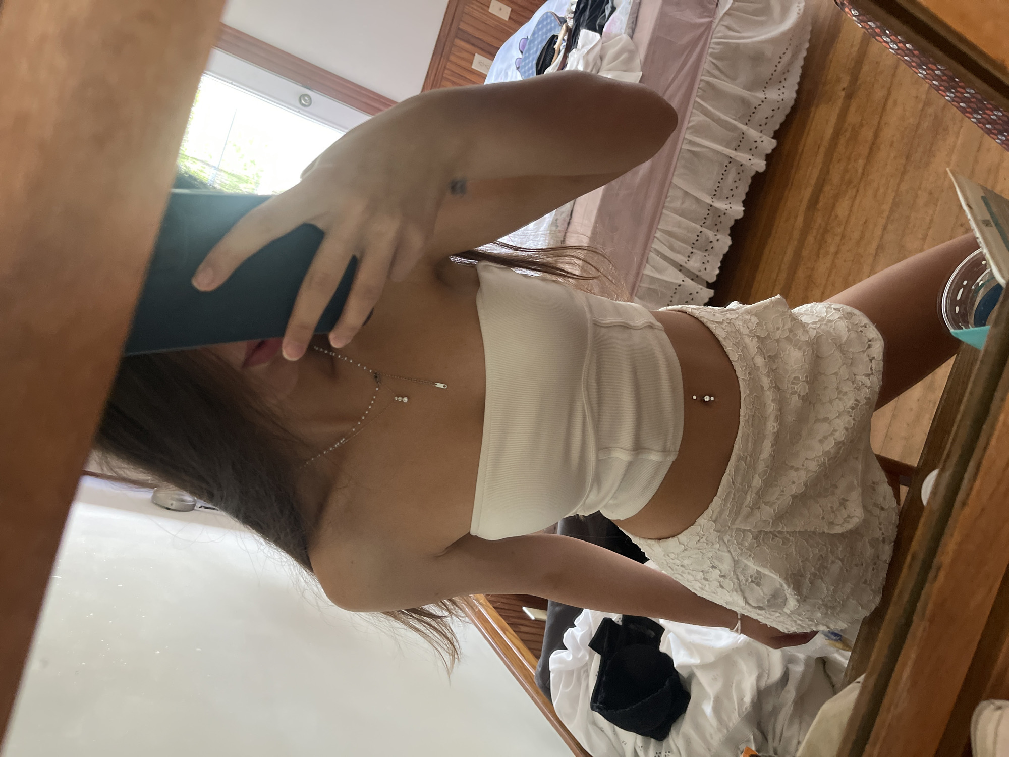 In this indoor, eye-level, vertical, medium shot, a person is taking a mirror selfie in what appears to be a bedroom. The subject is a young person with long, brown hair, wearing a white ribbed tank top and a white lace skirt. They are holding a dark-colored phone to take the photo, obscuring part of their face. A belly button piercing with two small gems is visible. The setting includes a wooden floor, a wooden headboard, and a window with natural light streaming through. A white ruffled bed skirt and some laundry are also visible in the background, suggesting a casual and lived-in environment. The overall mood is relaxed and possibly playful, typical of a personal selfie. There is no visible text in the image. The location context provided is Oak Bluffs, United States of America, which suggests a coastal or island setting, though this is not directly evident from the image itself. The lighting suggests it could be daytime.