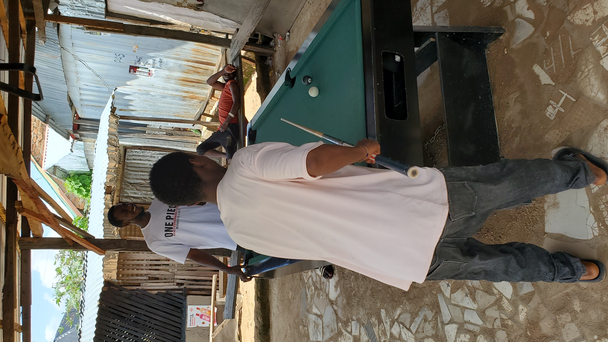 In Jalingo, Nigeria, an outdoor billiards scene unfolds. Two young men are engaged with a pool table. One man, wearing a white "ONE PIECE" t-shirt, stands to the left, observing. The other, in a light pink t-shirt and dark jeans, is actively playing, holding a pool cue over the green felt table. Two balls, one white and one black, are on the table. In the background, another person lies on a wooden bench. The environment appears somewhat rustic, with corrugated metal structures and wooden planks suggesting an informal setting. The ground is a mix of sand and broken tiles.