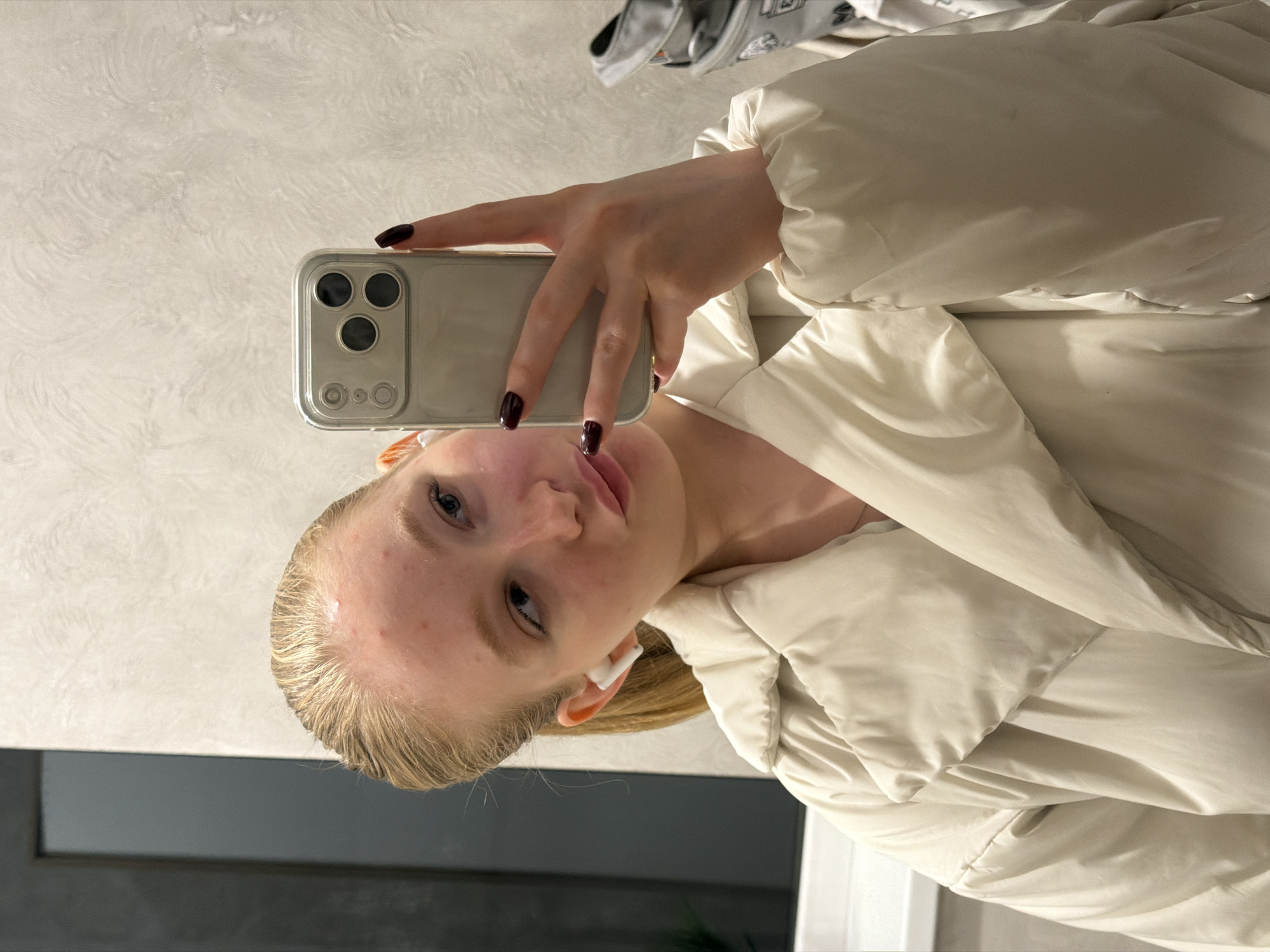 A woman is holding a smartphone in her right hand, taking a selfie. She has blonde hair, fair skin, and is wearing a white puffer jacket. The phone has a reflective case. The location appears to be indoors, with a gray wall and a glimpse of light near the bottom, but it is not possible to determine the location within the city of Rossiyskiy, Russia.